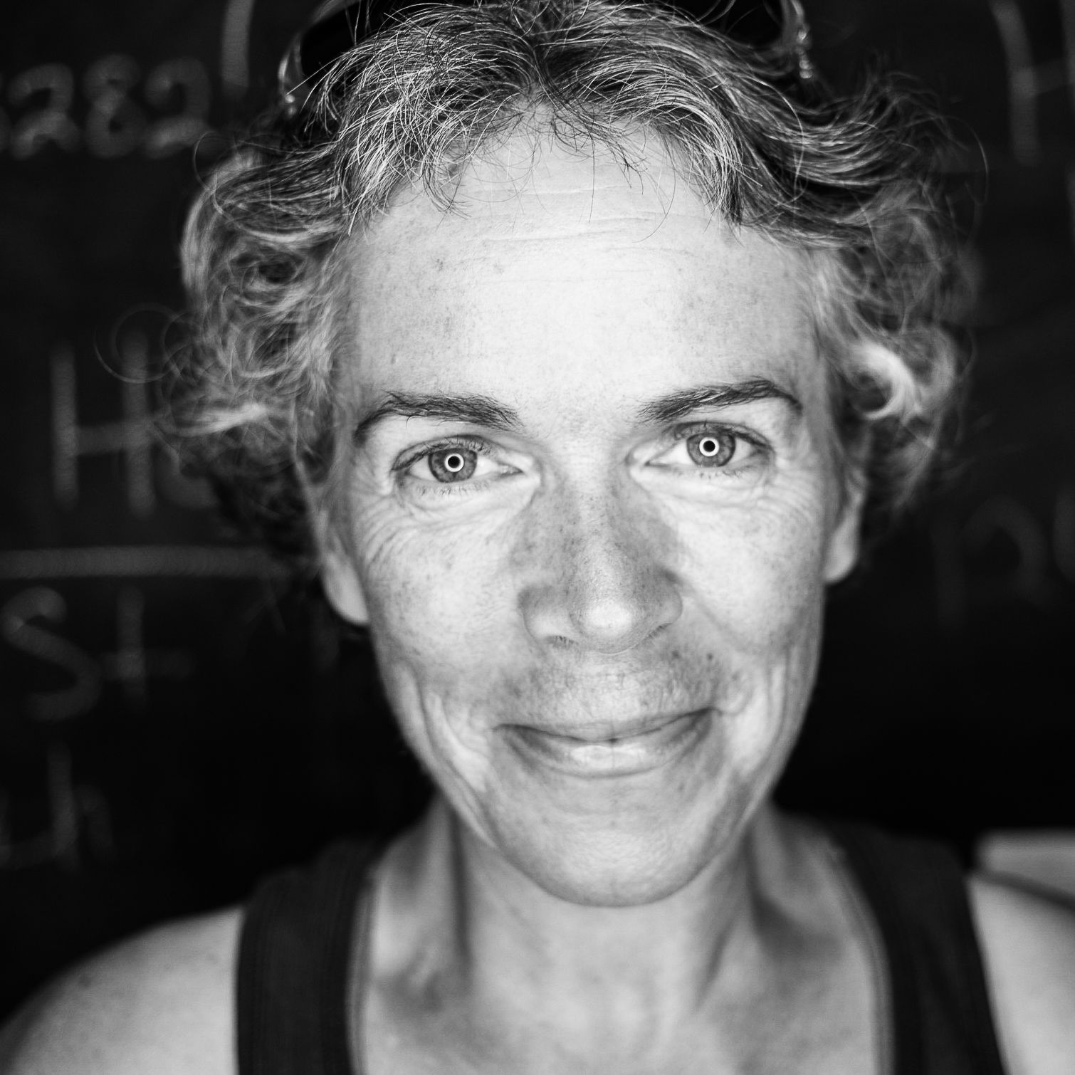 Woman with curly gray hair smiles at the camera, wearing a dark top, with chalkboard background.