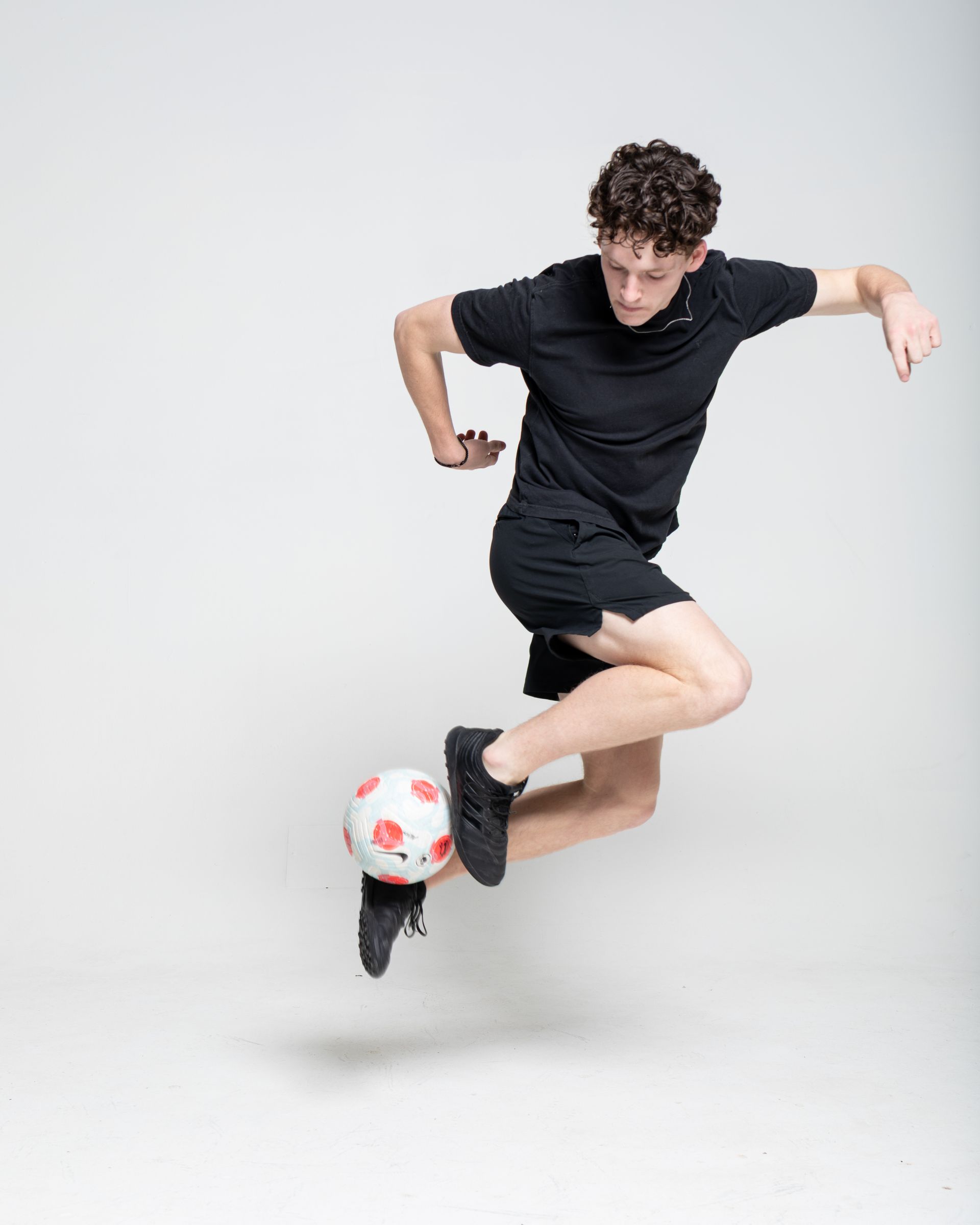 Person in black clothing leaping, juggling soccer ball with foot against white backdrop.
