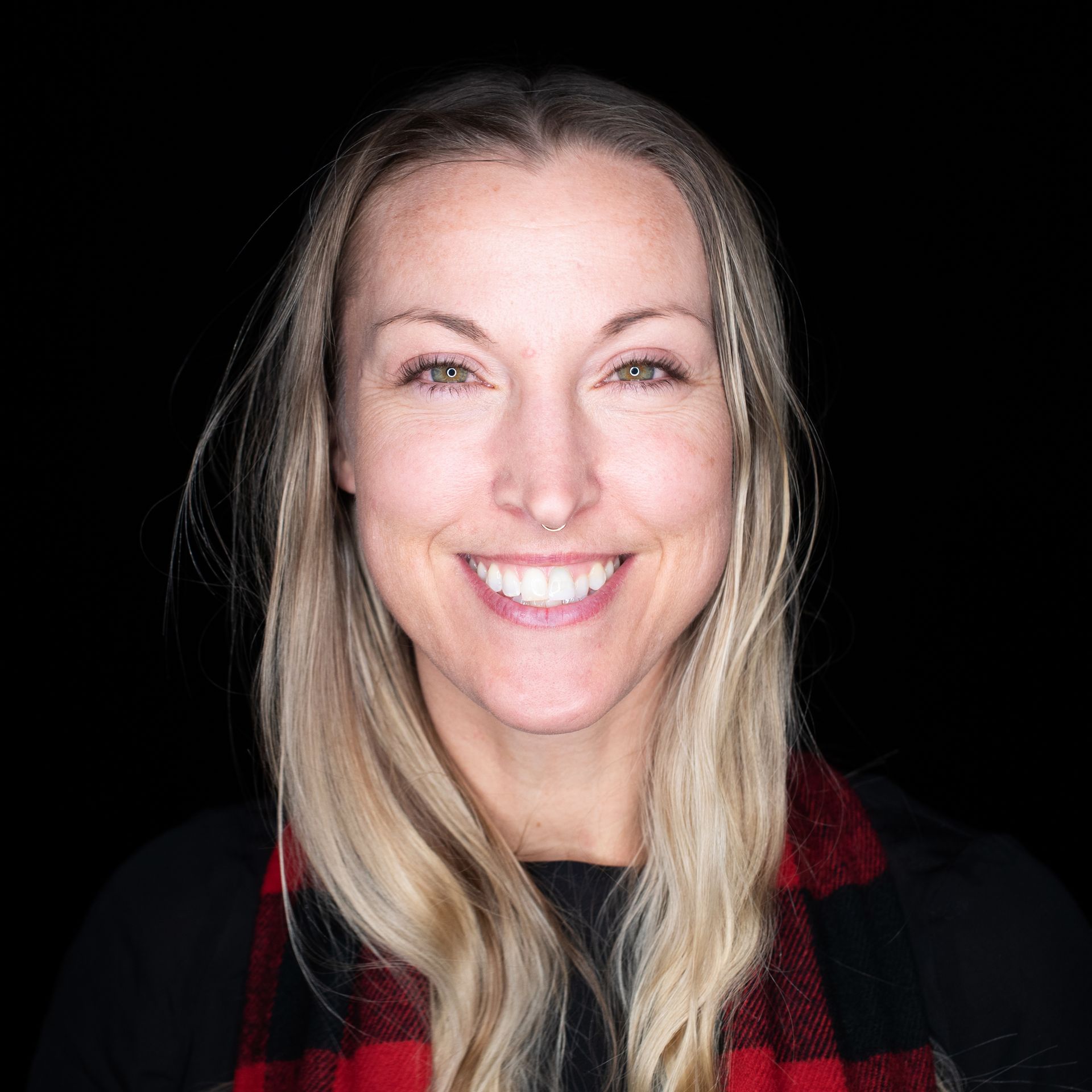 Woman with long blonde hair, smiling at the camera, wearing a red and black scarf. Black background.