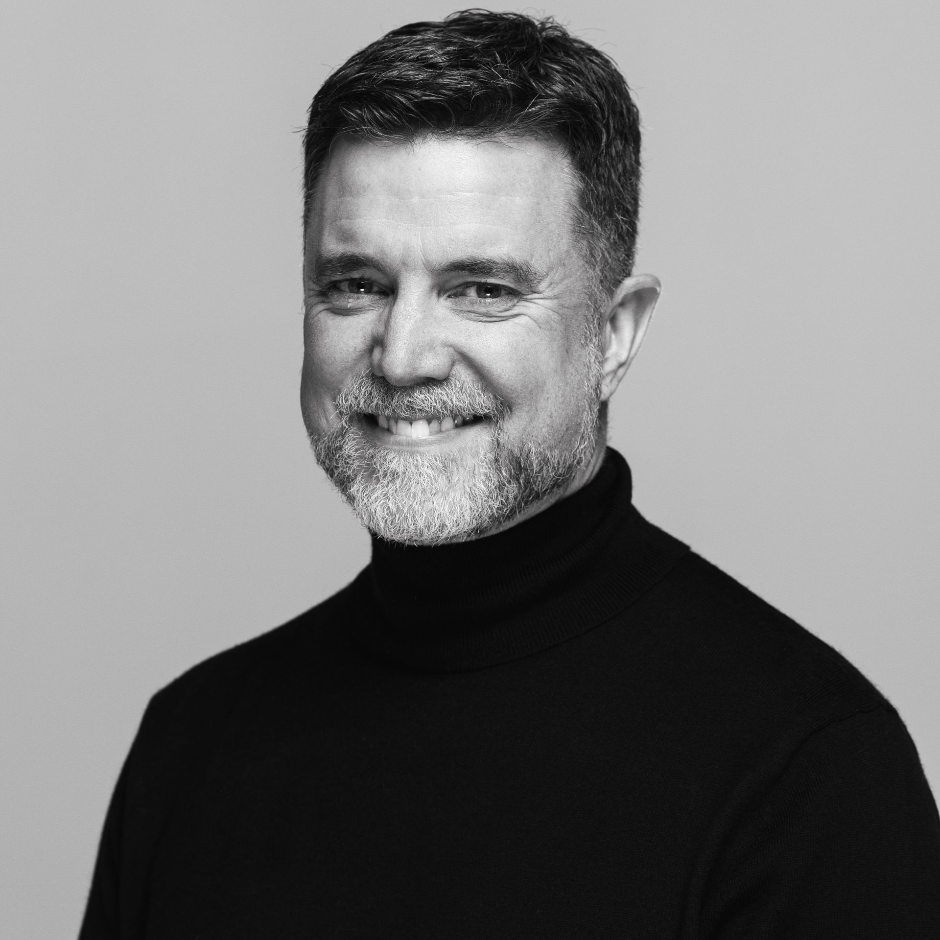 Man in black turtleneck smiles, graying beard and short hair. Studio background.