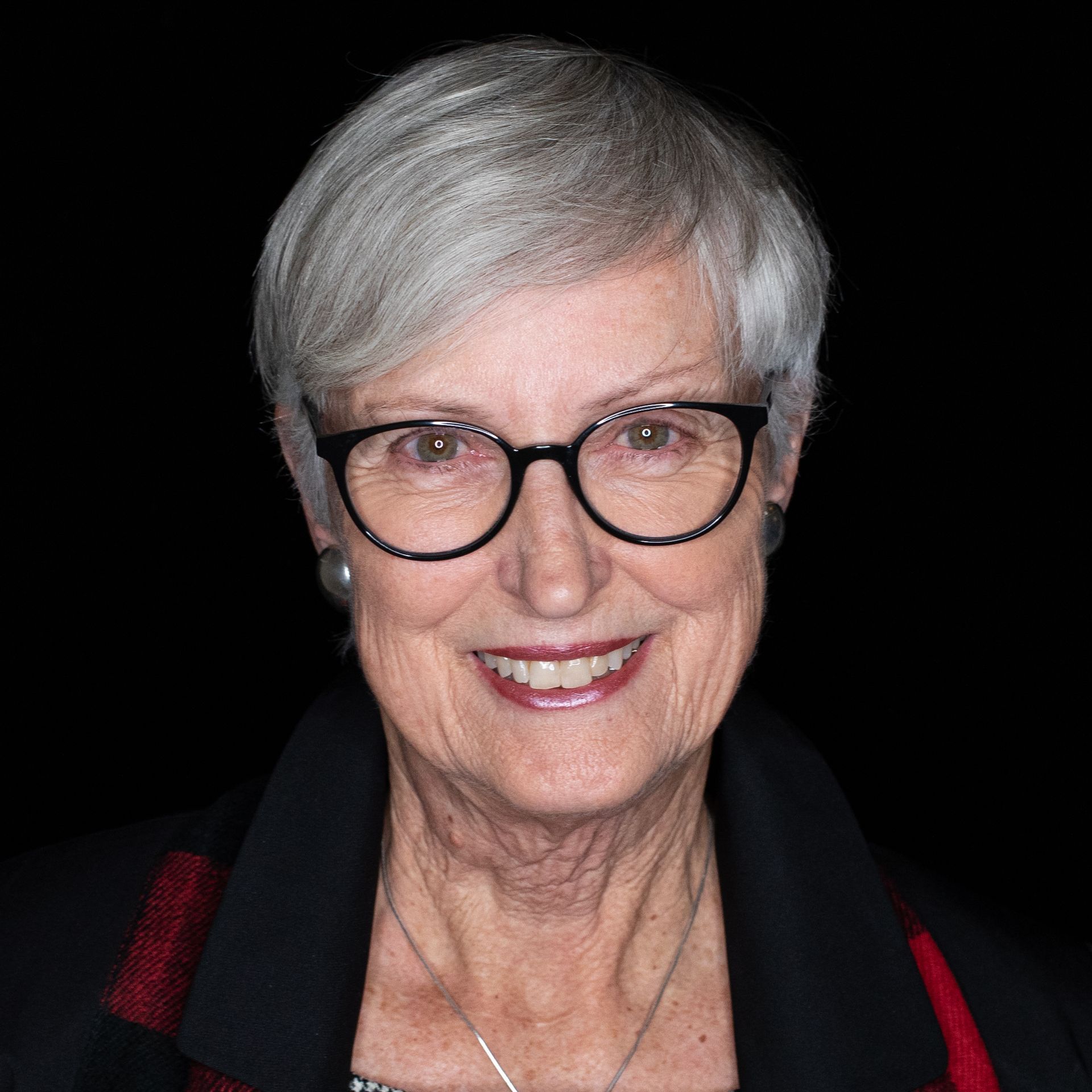 Smiling woman with short gray hair and glasses, wearing a black blazer with a red collar, against a black background.