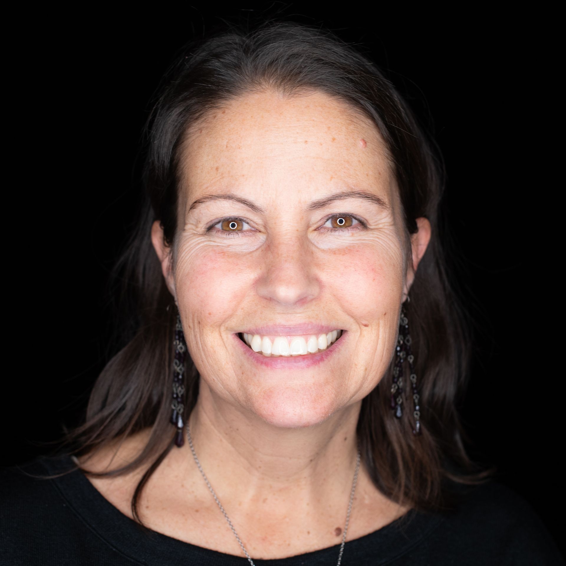 Woman with brown hair smiling, wearing a black shirt, earrings, and necklace, against a black background.