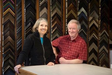 A man and a woman are posing for a picture in front of a wall of picture frames.