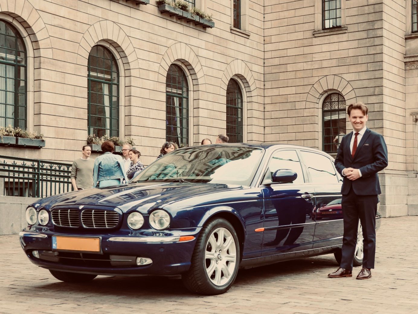 Een man in pak staat naast een donkerblauwe Jaguar voor een stenen gebouw met boogvensters.