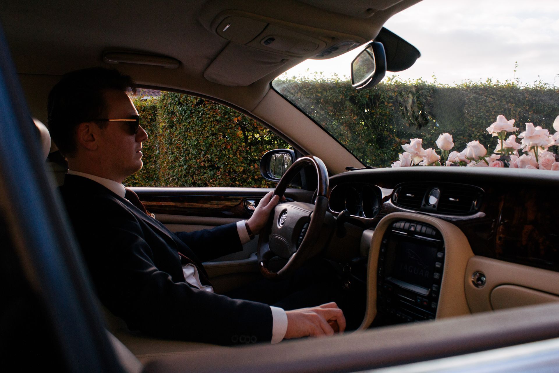 Man met zonnebril rijdt in een luxe auto, armen aan het stuur, beige interieur.