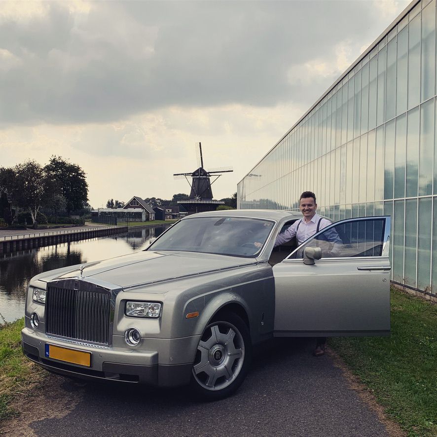 Een man glimlacht naast een zilveren Rolls-Royce. Op de achtergrond, vlakbij een waterweg, zijn een windmolen en een glazen gebouw te zien.