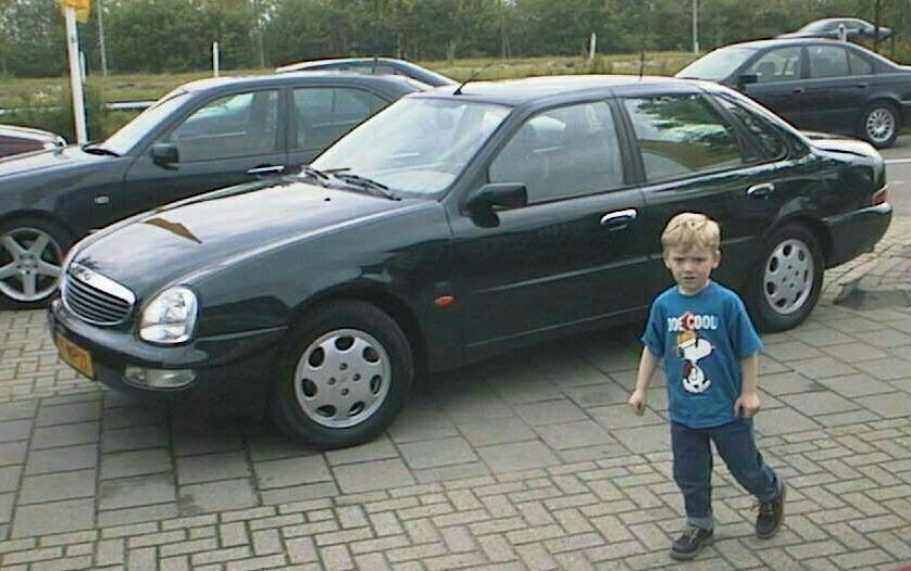 Een donkergroene sedan staat geparkeerd, een klein jongetje in een blauw shirt loopt voorbij.