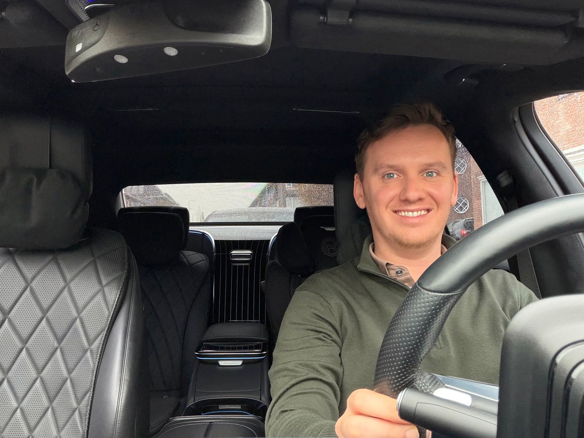 Een glimlachende man houdt het stuur vast in de auto en kijkt naar de camera.