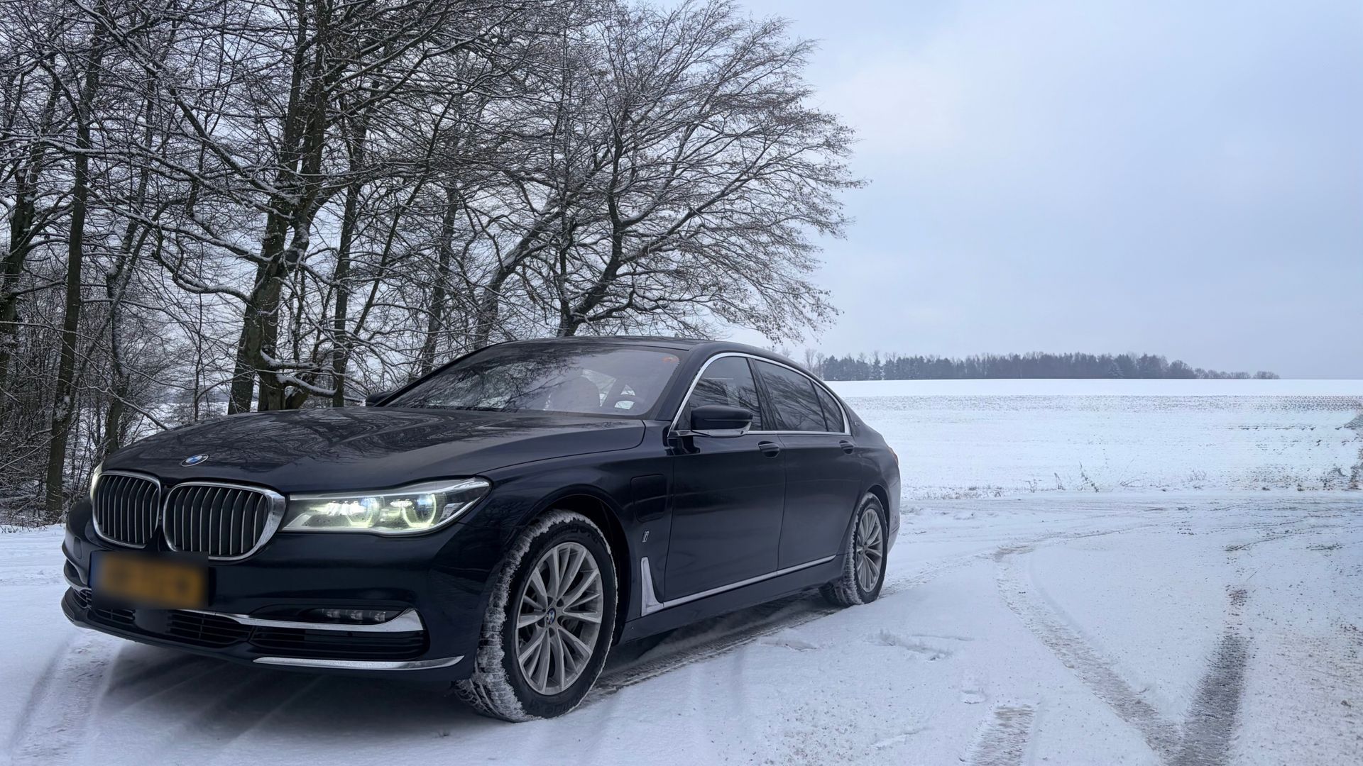 Een donkerblauwe BMW sedan staat geparkeerd in een met sneeuw bedekt veld bij bomen op een bewolkte dag.