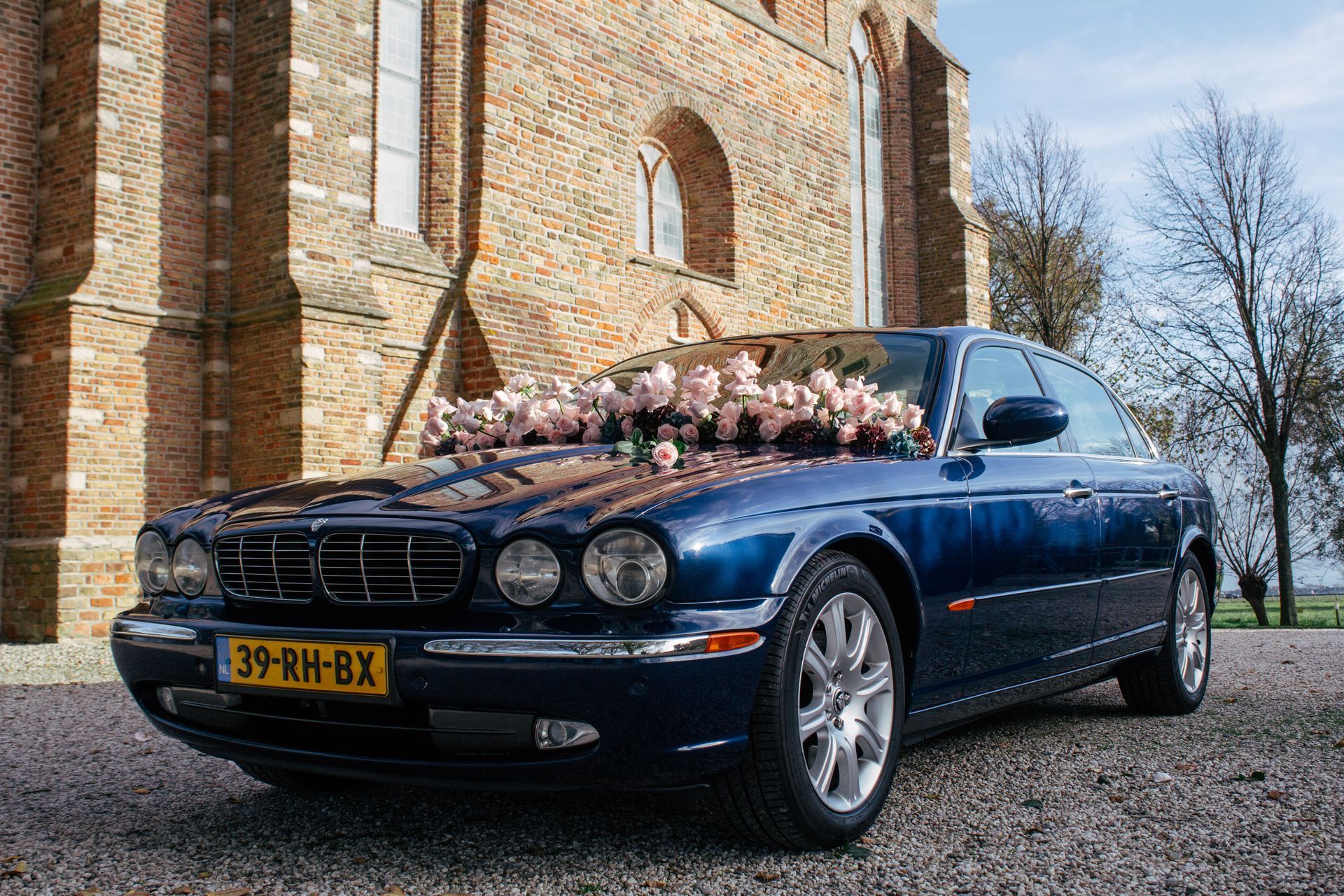 Een blauwe Jaguar met bloemversiering staat geparkeerd voor een bakstenen gebouw.