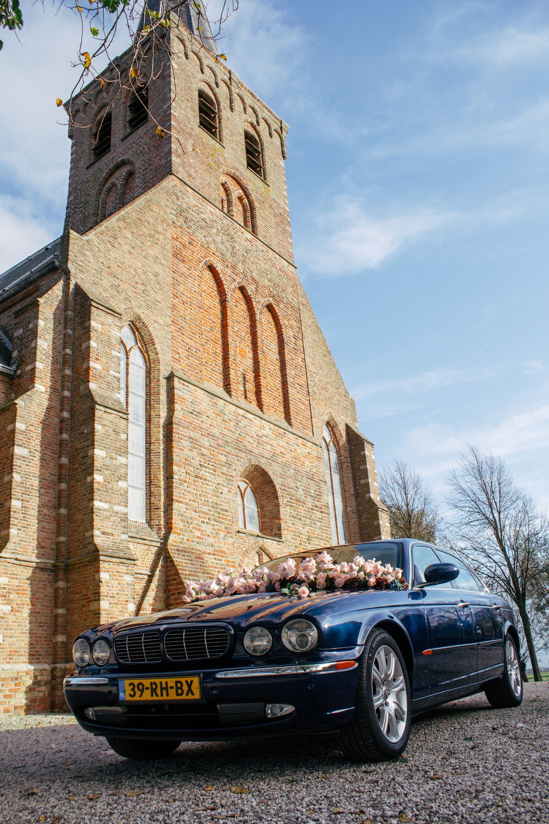 Een blauwe jaguar met bloemversiering staat geparkeerd voor een bakstenen kerk op een zonnige dag.