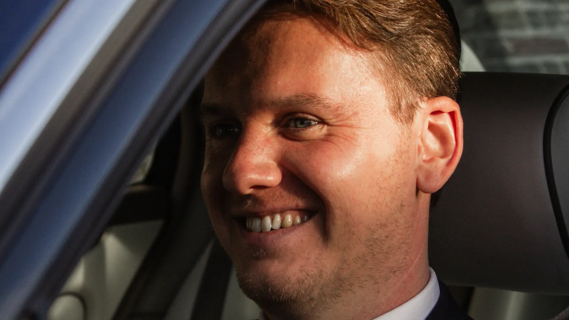 Een man glimlacht in een auto, verlicht door de zon.