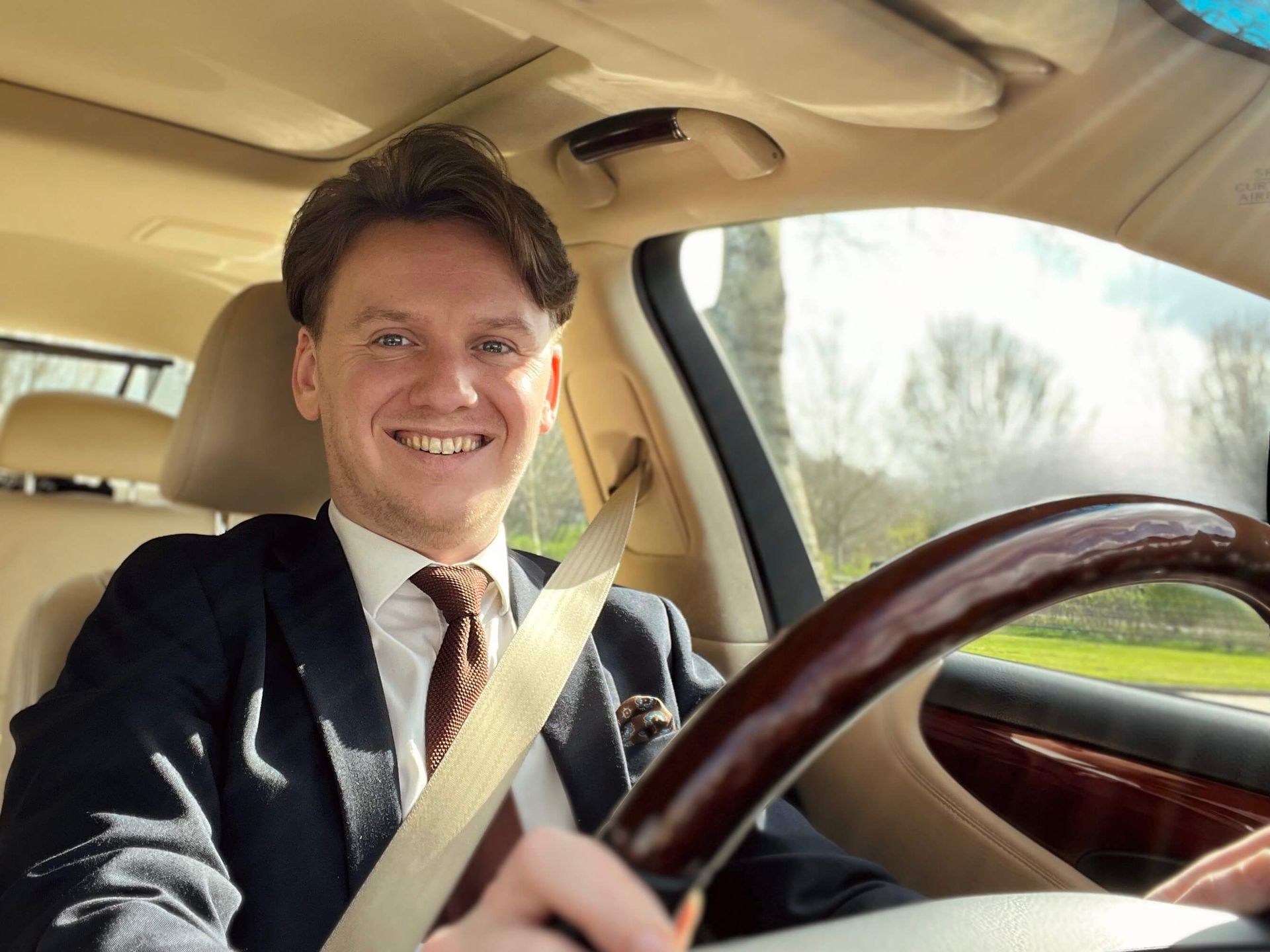 Een lachende man, rijdend in een auto, gekleed in pak en stropdas.