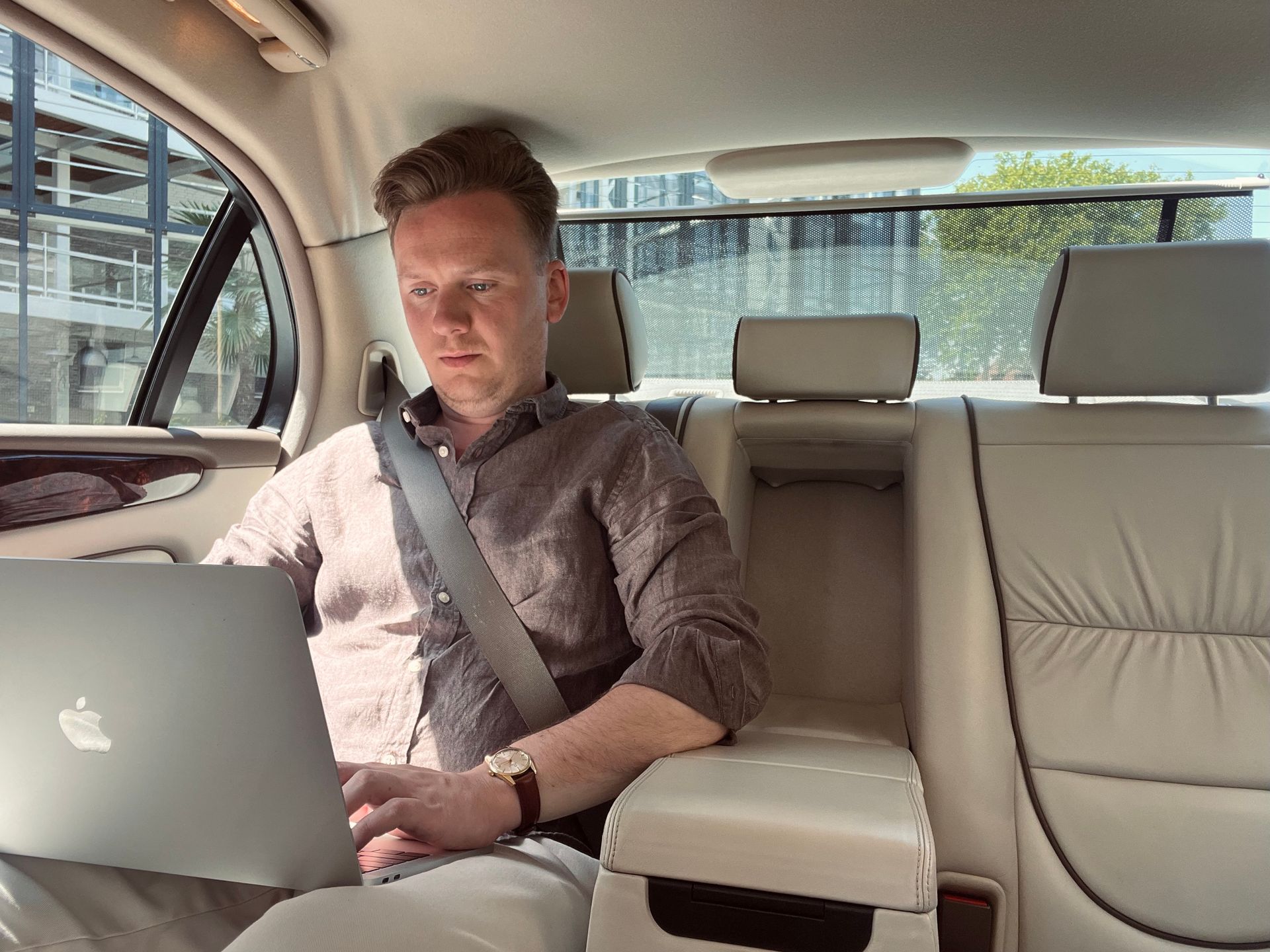 Man in een auto, met veiligheidsgordel om en werkend op een laptop. Interieurfoto met beige lederen stoelen.