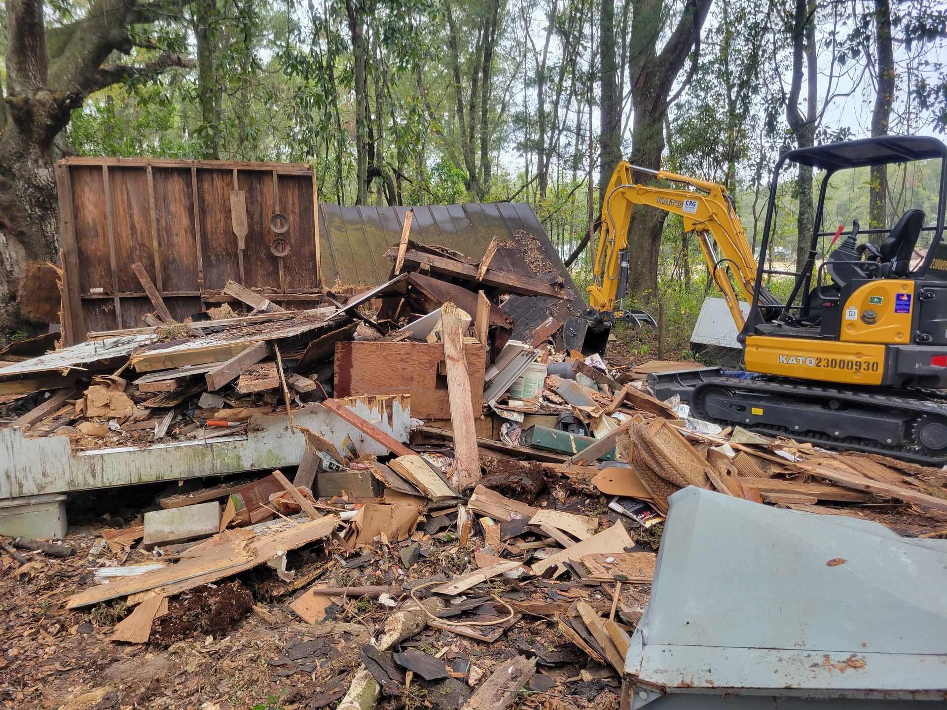 Demolition of a wooden structure, debris pile with a yellow excavator in a wooded area.