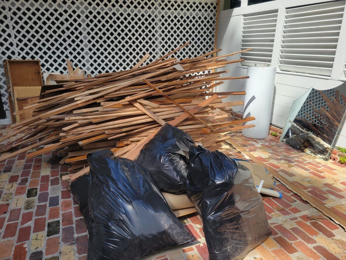 Wood debris pile with black trash bags on a brick patio near a white window.