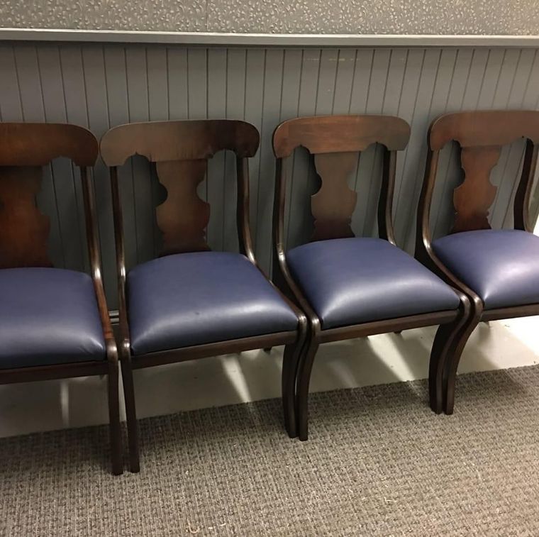 Four empty wooden chairs with blue seats lined up against a paneled wall in a room.