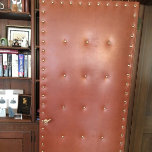 Padded brown door with brass studs and a small handle, set beside a wooden bookshelf.