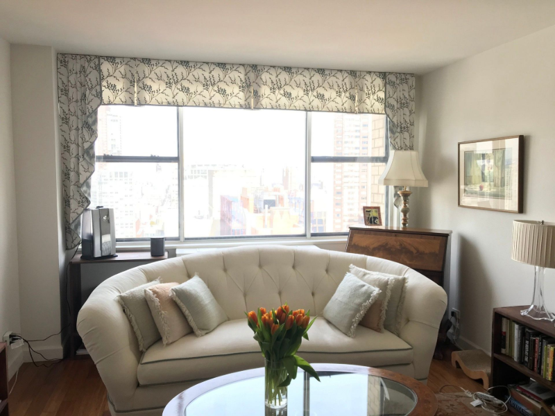 Bright living room with a cream sofa, large window, flowers on a table, and a lamp and piano in the corner