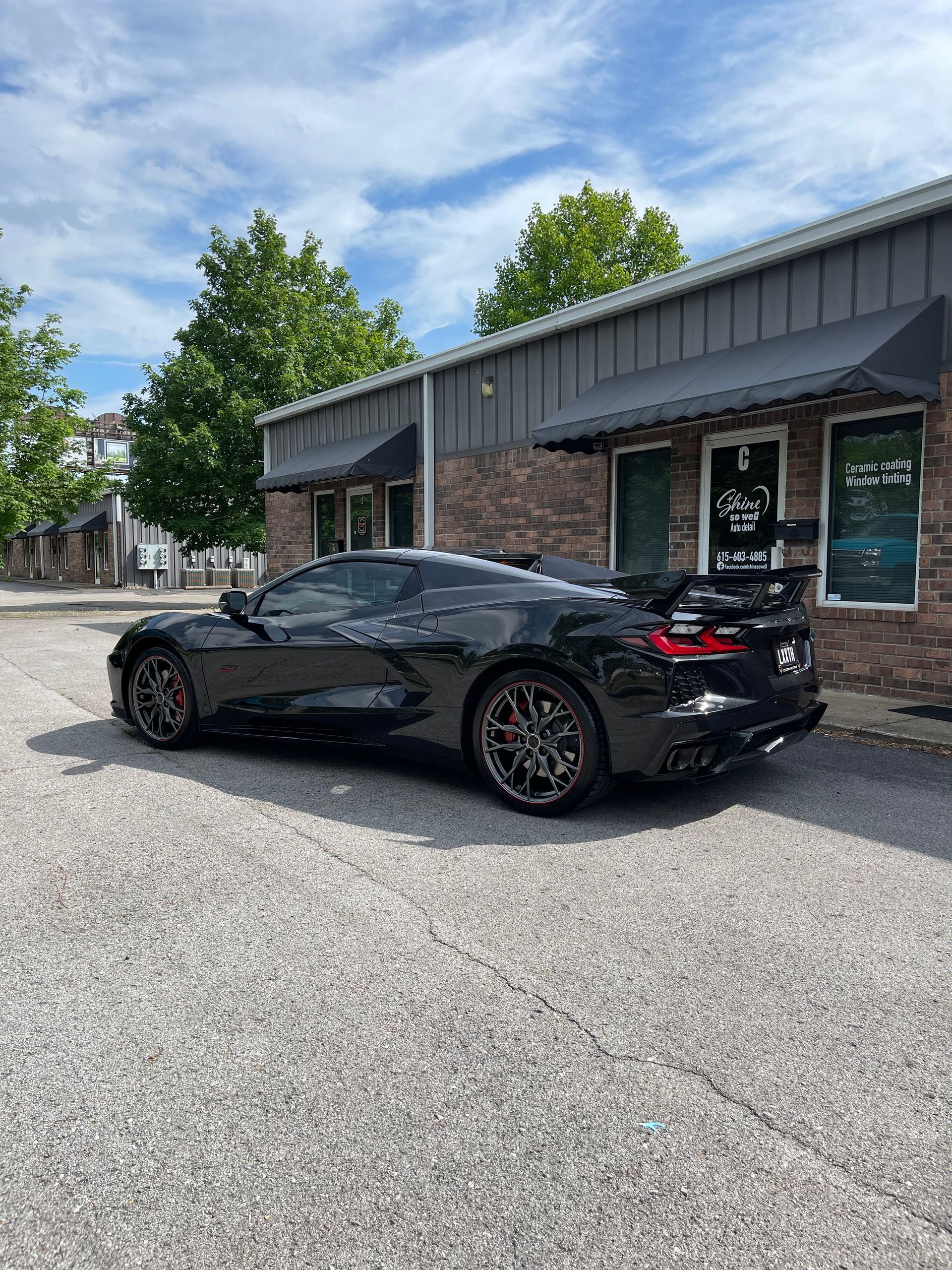 Black Corvette sports car parked in front of a building with awnings. Gravel driveway, partly cloudy sky.