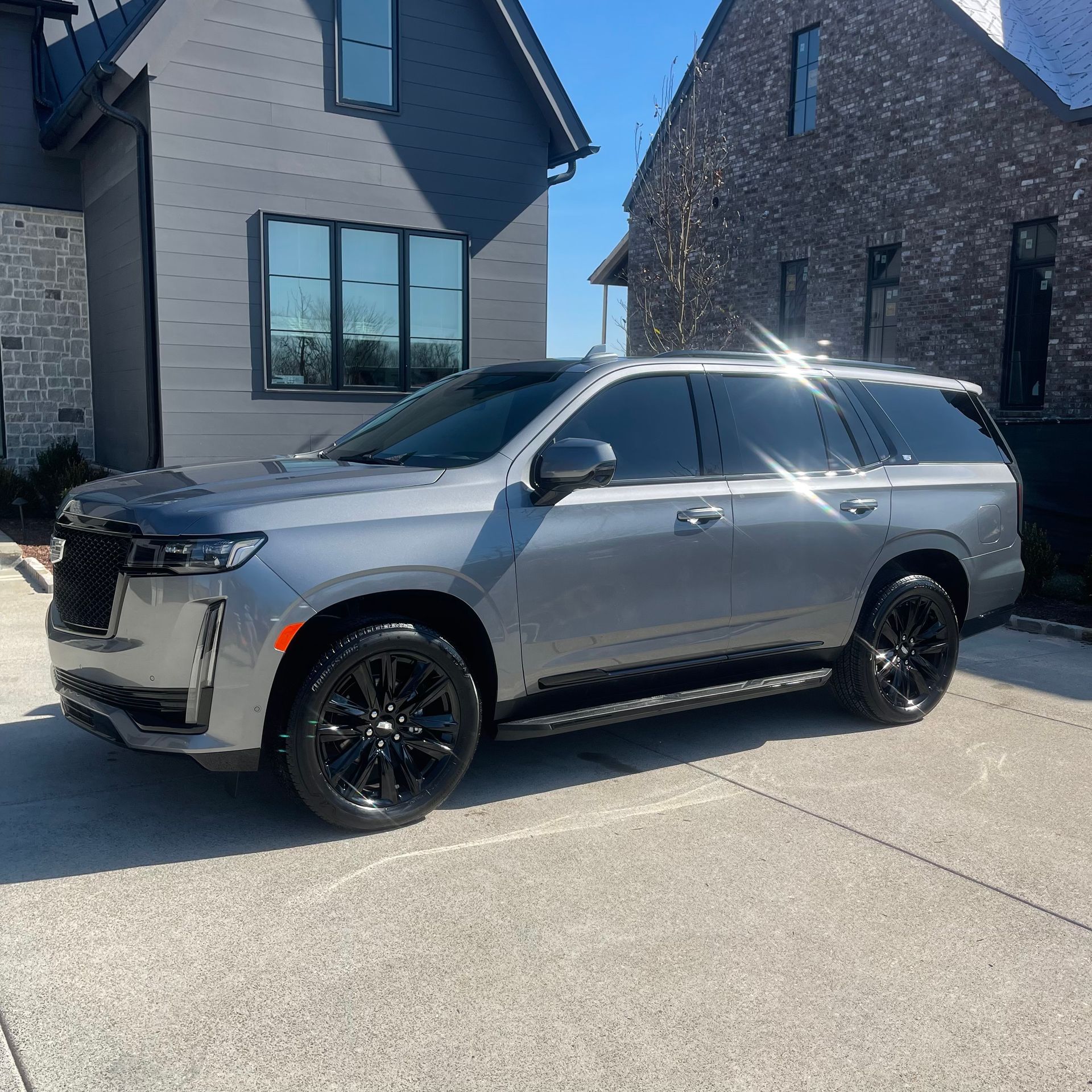 Gray Cadillac Escalade SUV parked in front of a modern gray and brick house with black wheels and tinted windows.