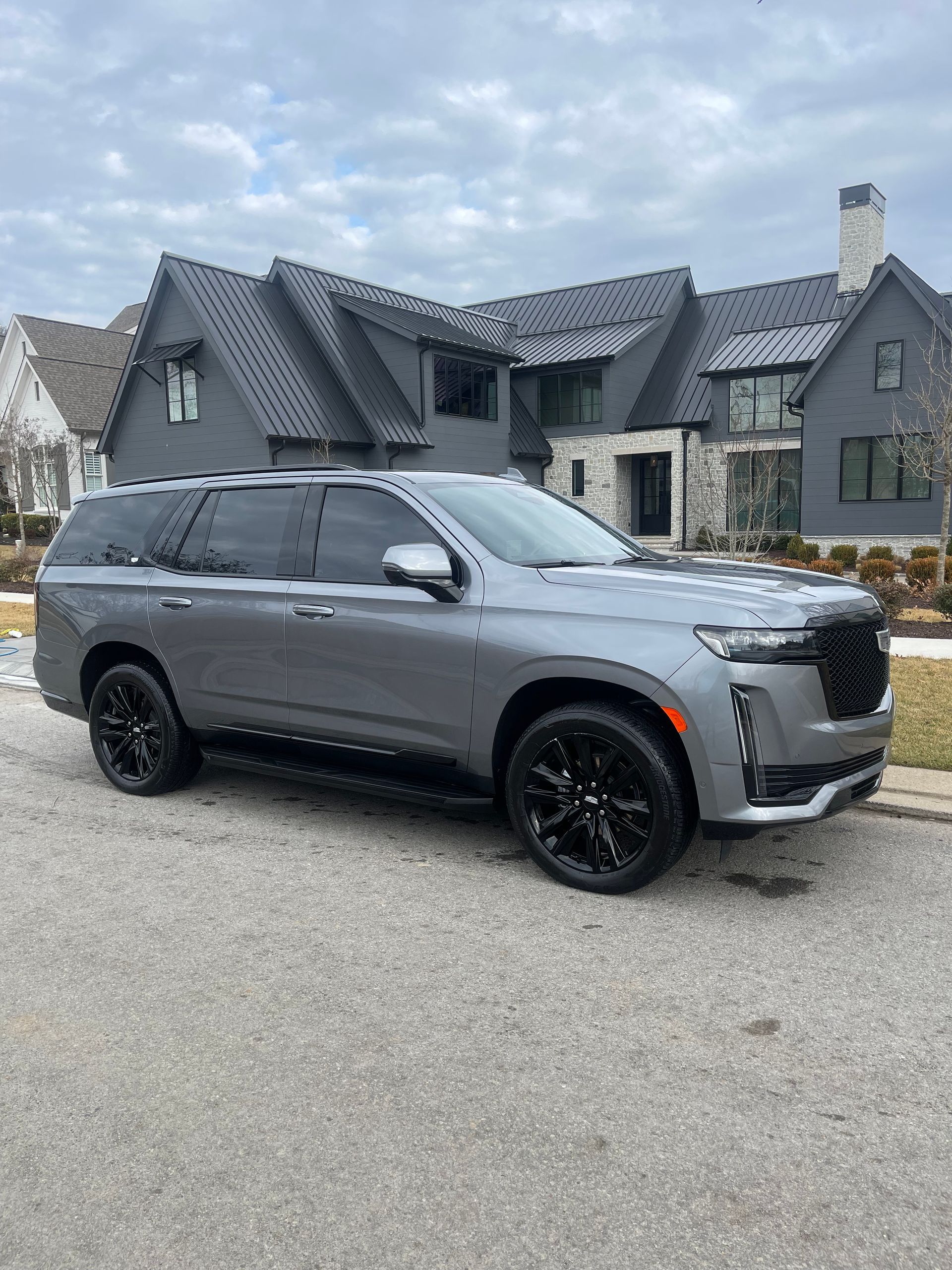 Gray Cadillac Escalade parked on a paved road in front of a modern gray house. Black wheels. Cloudy sky.