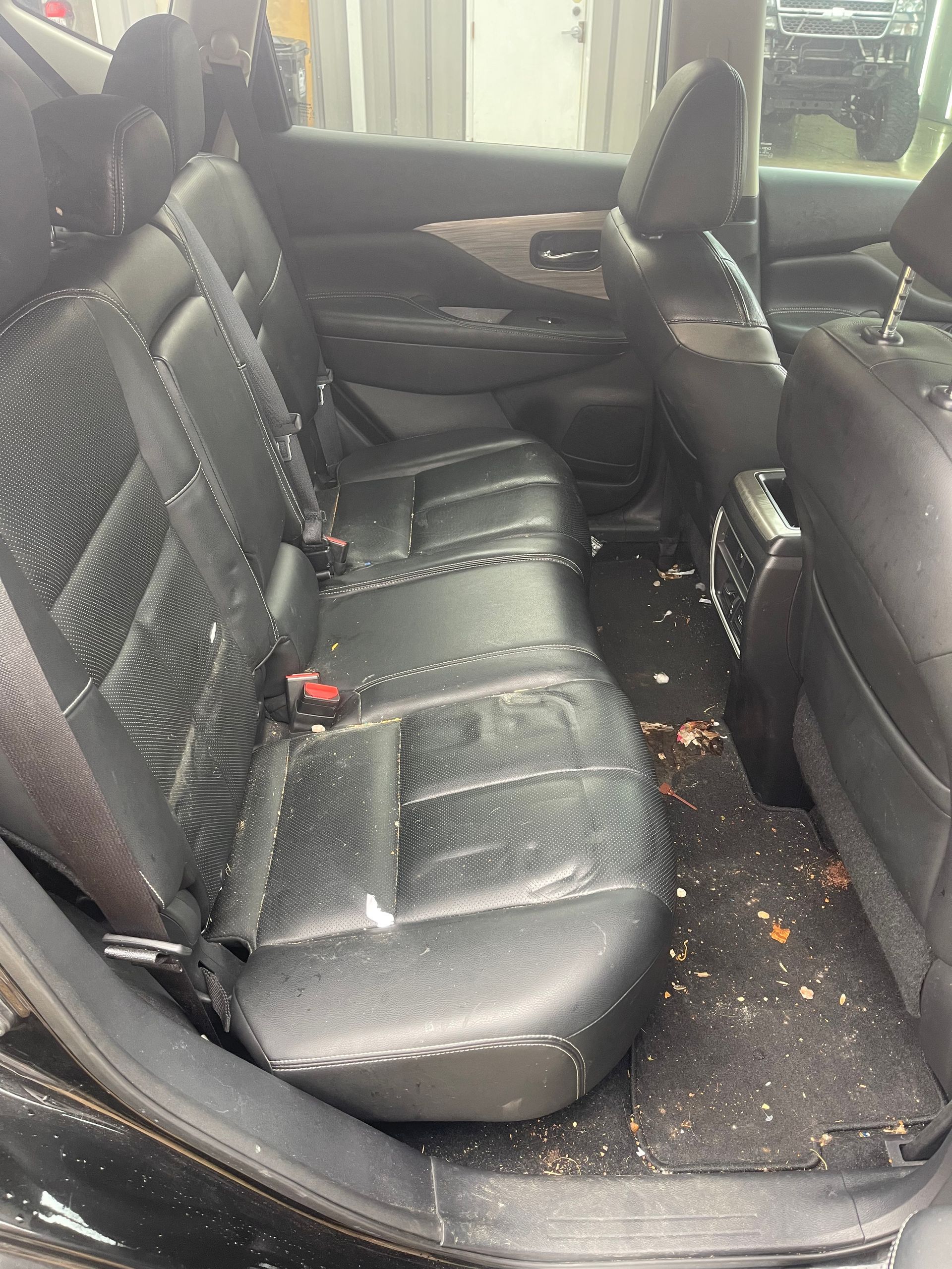 Back seat of a black car, dirty with debris on seats and floor. Leather seats.