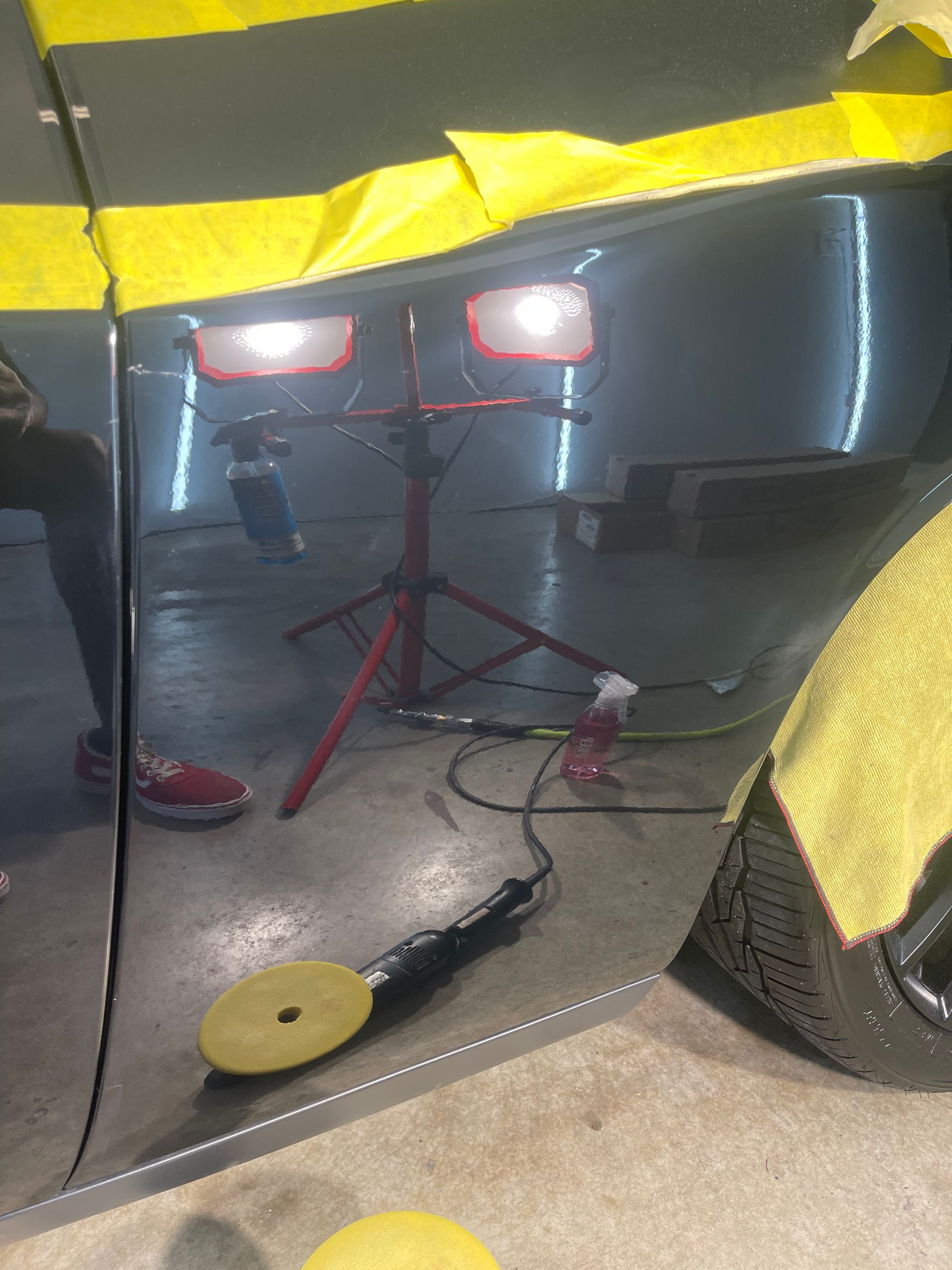 Car door being polished with a buffer, reflected in a shiny surface. Yellow tape and detailing lights are visible.