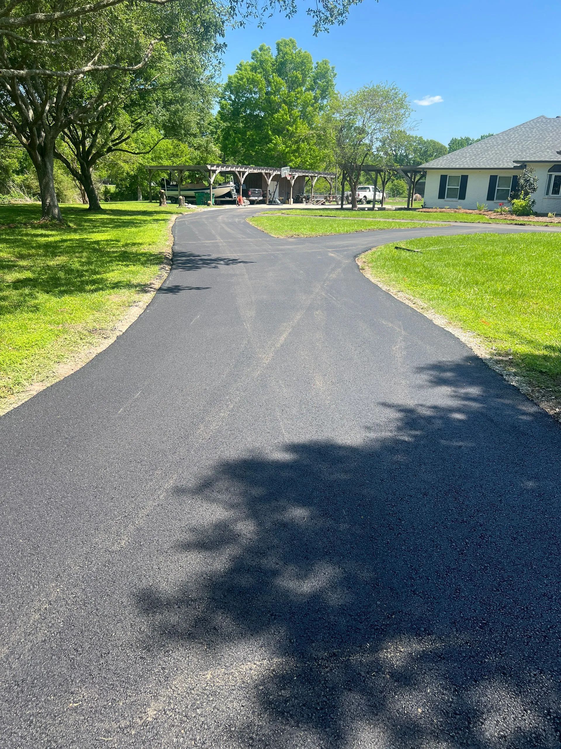 Dark asphalt driveway splits, leading to a house and a shaded structure on a sunny day.