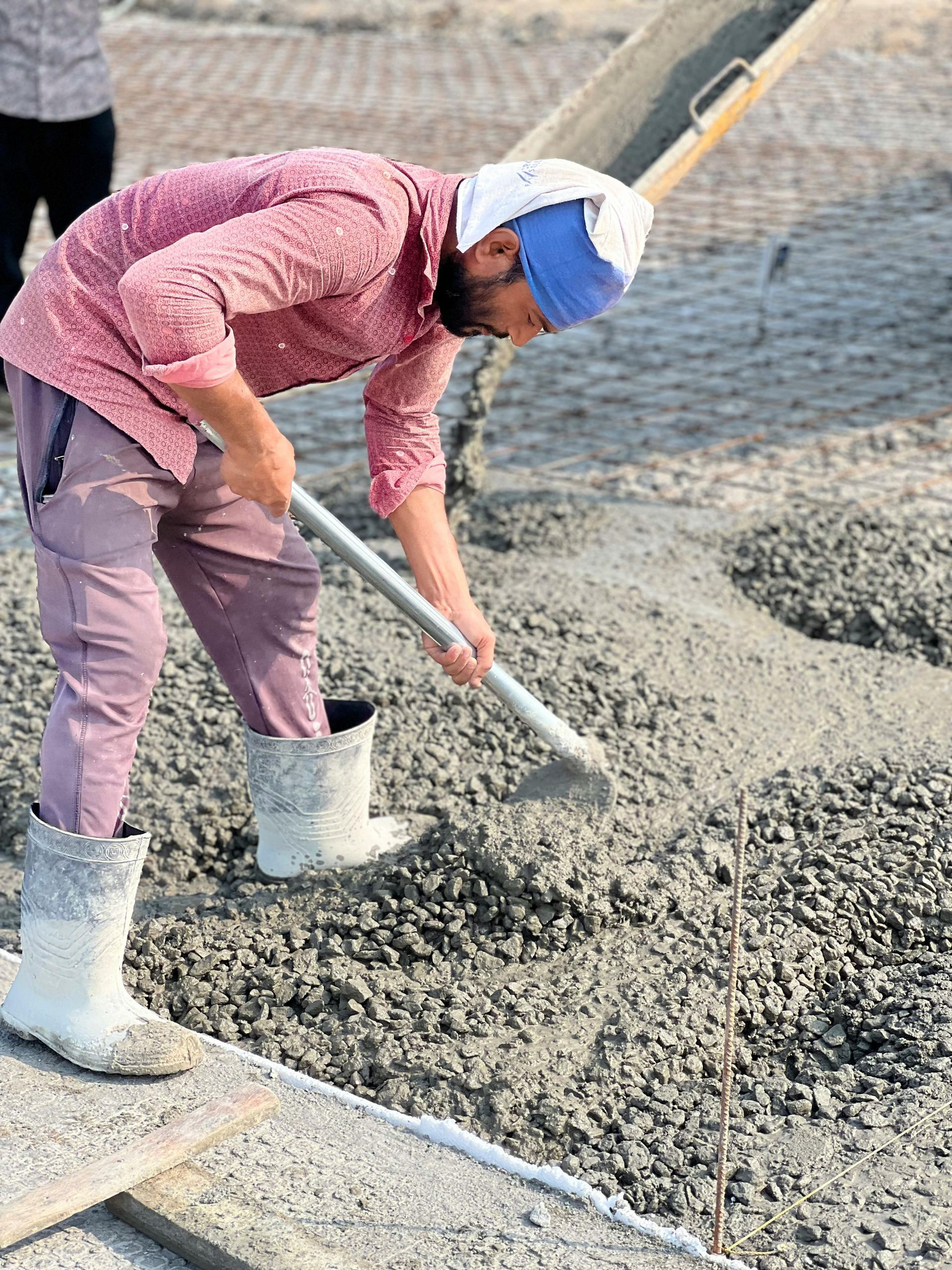 Construction worker shoveling concrete from a chute, wearing boots and a head covering, outdoors.