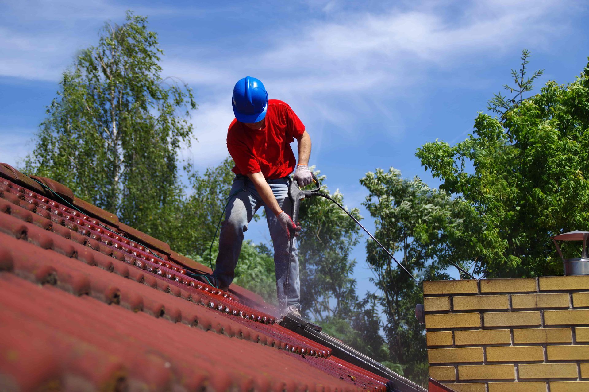 Person wearing a blue hard hat and red shirt power washing a red tile roof.