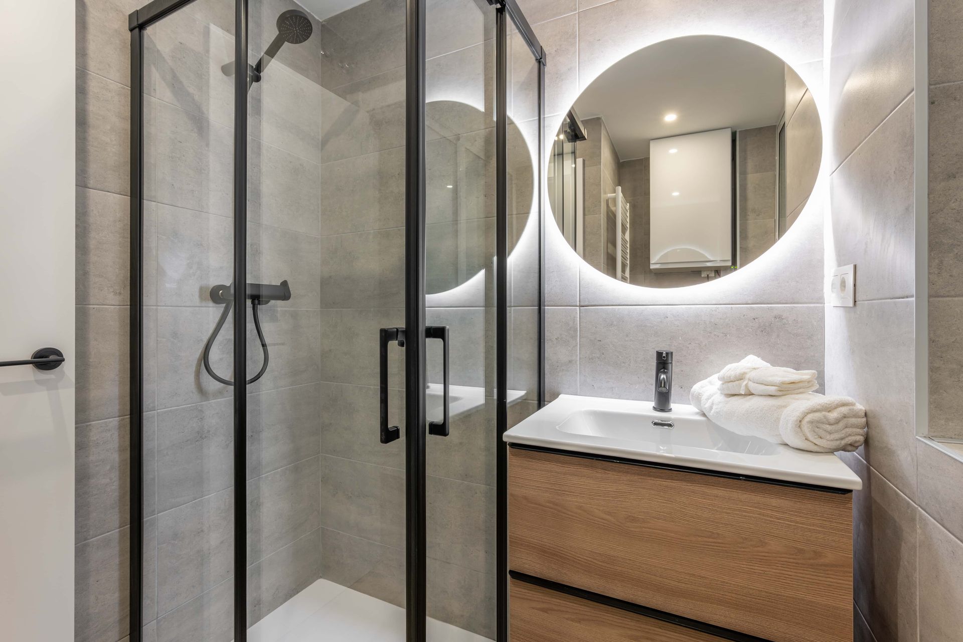 Bathroom with a glass shower, wooden vanity, round mirror, and gray tile.