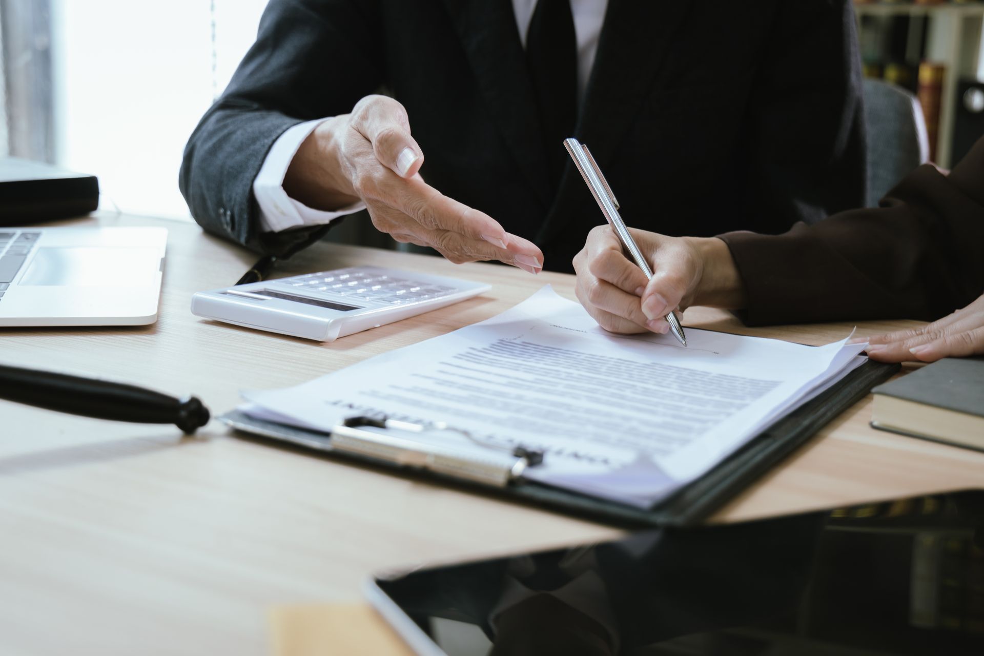 Person signing a legal document while a professional reviews paperwork across a desk. Person signing a legal document while a professional reviews paperwork across a desk.