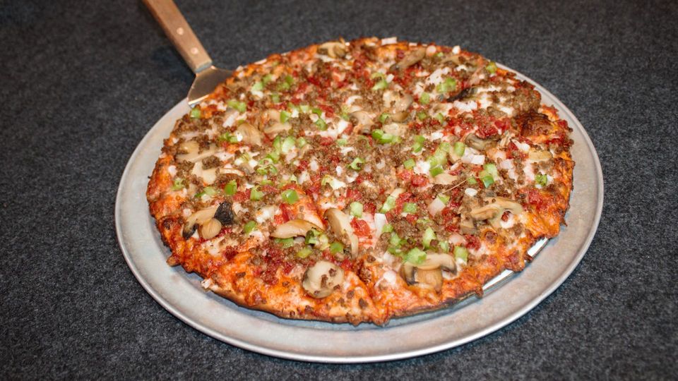 Large loaded pizza on a metal tray with a wooden pizza peel handle visible on the left.