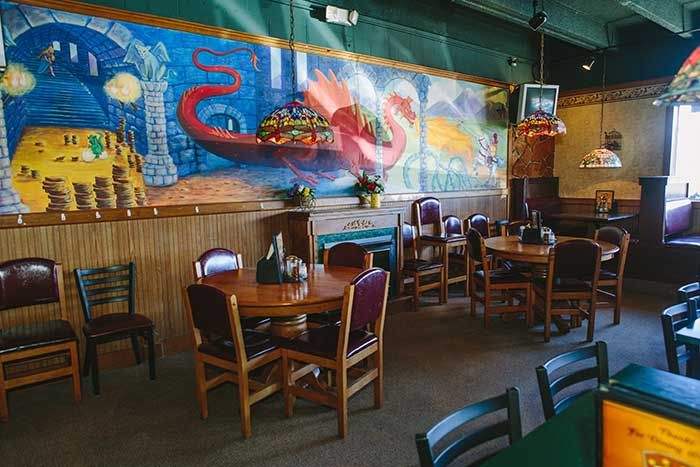 Empty restaurant dining area with wooden tables, chairs, and a colorful wall mural of a dragon and village scene