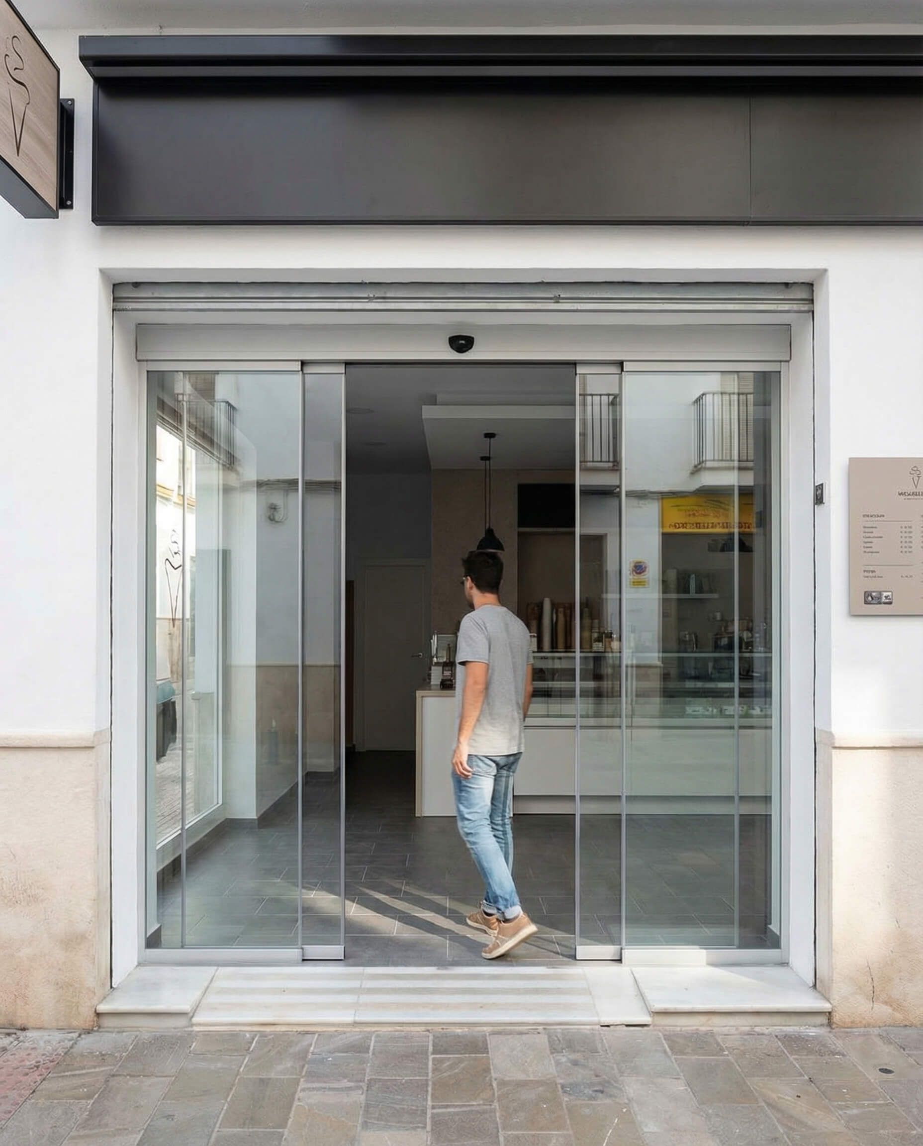 Hombre entrando en una tienda con puertas corredizas de cristal. El exterior del edificio es blanco con un cartel negro sobre la entrada.