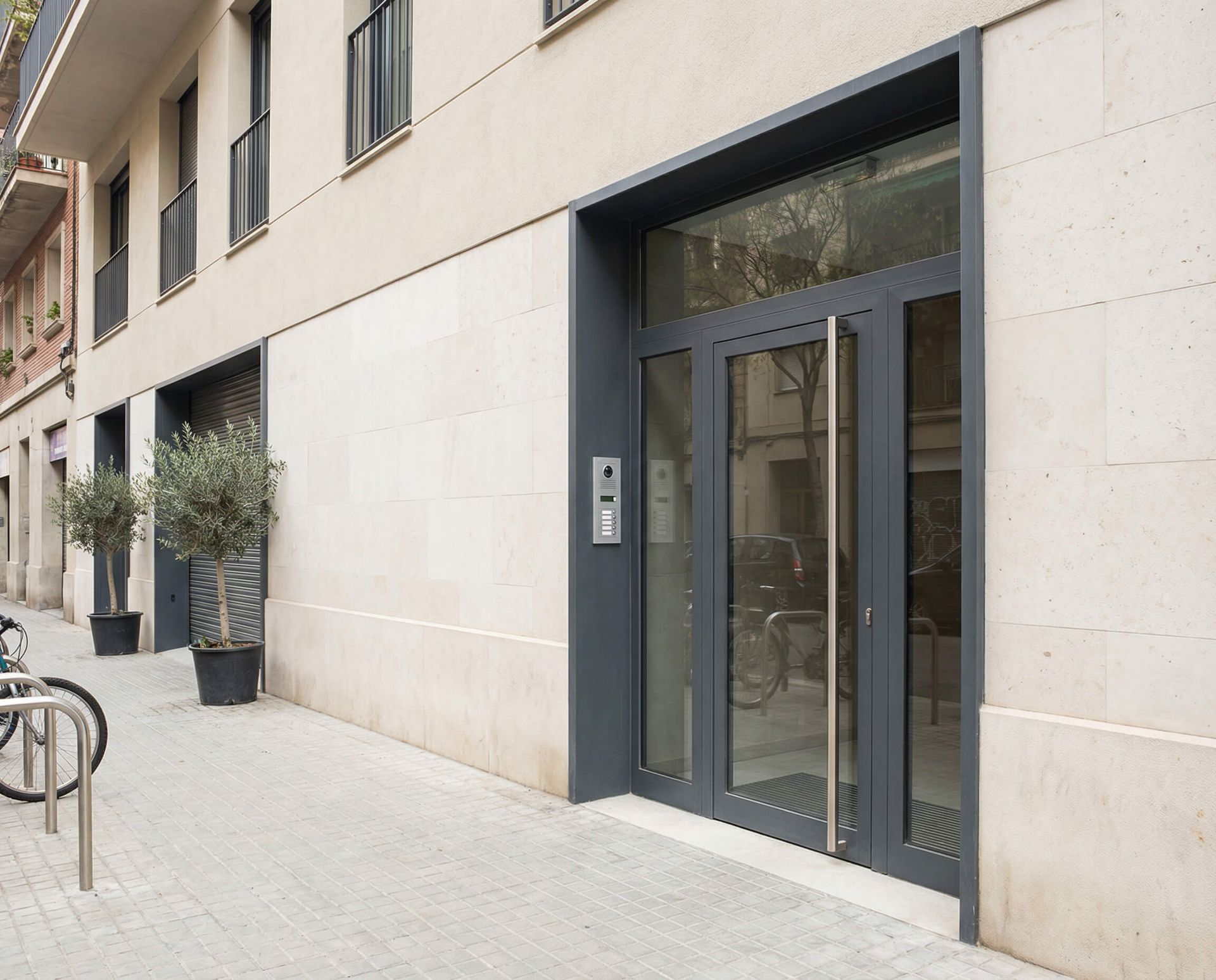 Entrada a un edificio con marco de puerta gris y puerta de vidrio, árboles en macetas y portabicicletas.