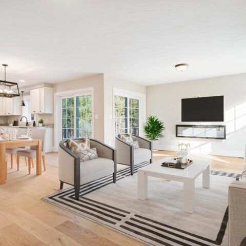 Bright open living room with gray sofas, striped rug, white coffee table, TV, and kitchen/dining area beyond