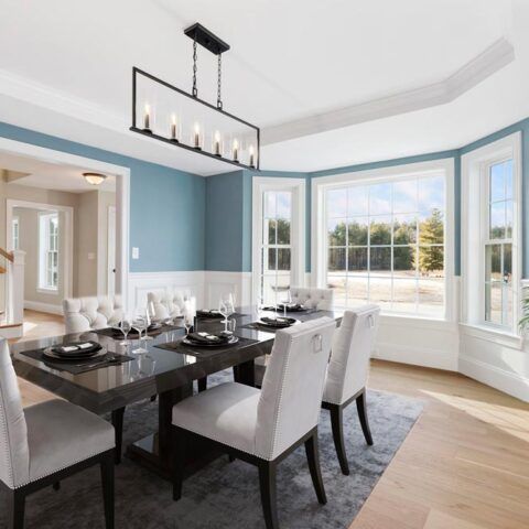 Modern dining room with dark table, white chairs, blue walls, and large bay windows overlooking greenery