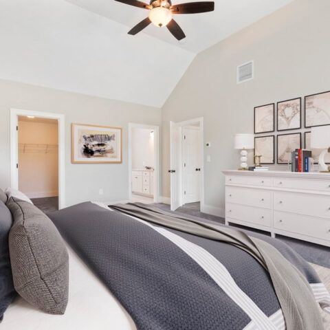 Bright bedroom with gray bedding, white dresser, framed wall art, and a ceiling fan.
