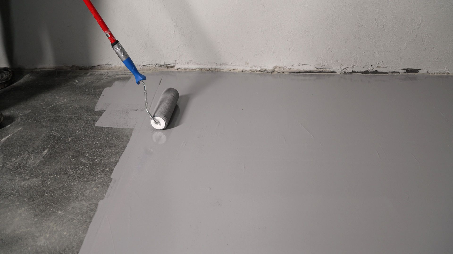 Une personne applique un enduit gris au rouleau sur un sol, recouvrant une surface en béton foncé.