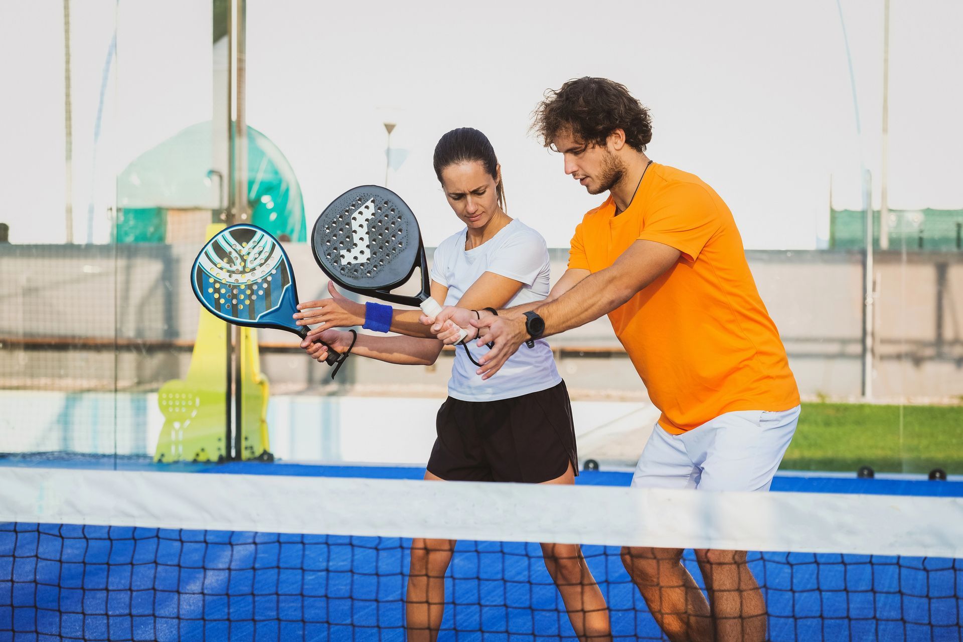 Un uomo insegna a una donna a giocare a padel, entrambi con le racchette in mano, vicino alla rete, all'aperto.