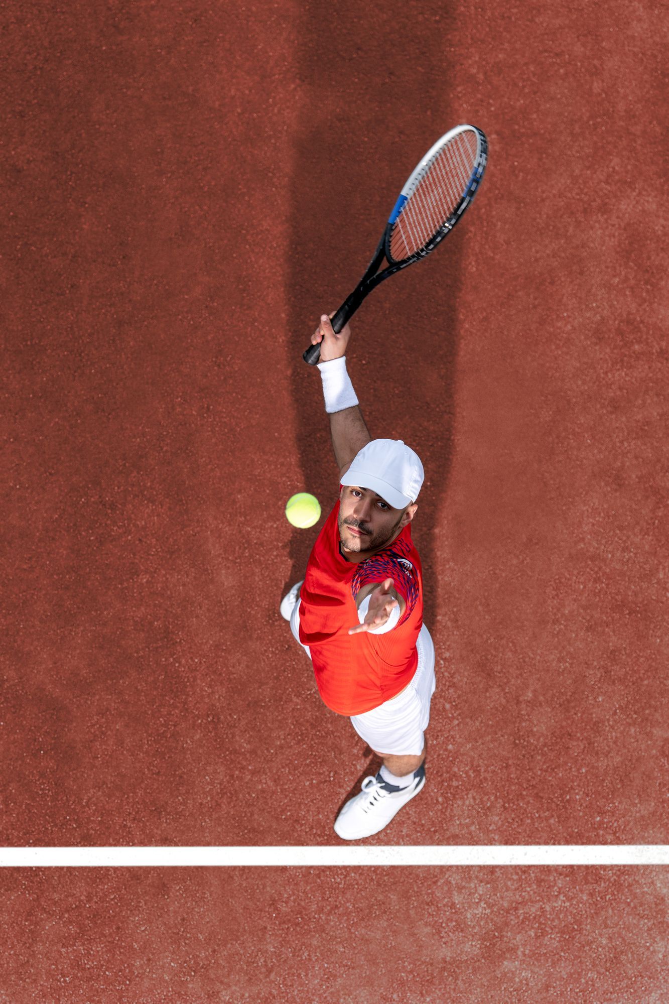 Giocatore di tennis che serve la palla, visto dall'alto, su un campo rosso.