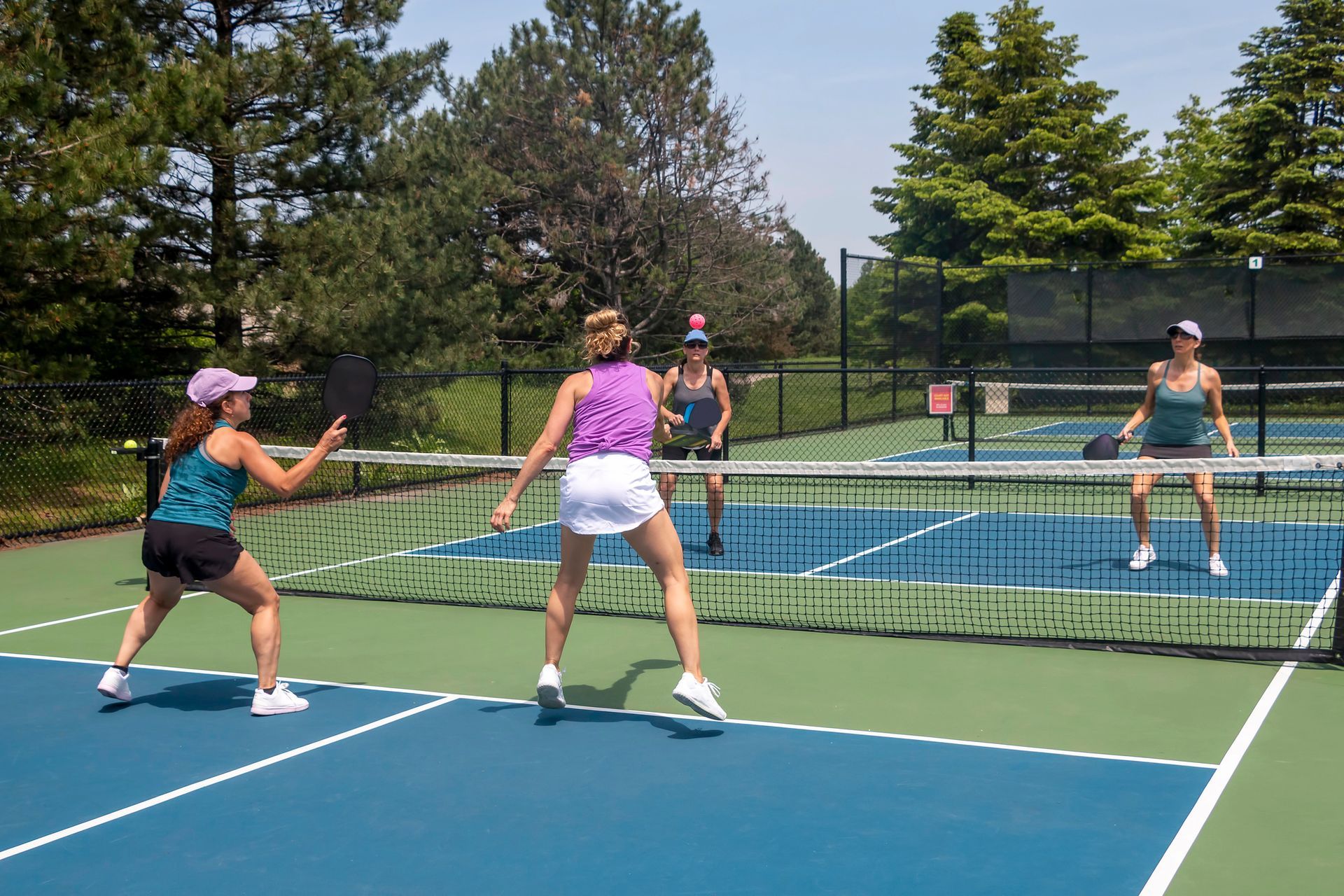 Quattro persone giocano a pickleball su un campo blu con rete verde, in un ambiente soleggiato all'aperto.