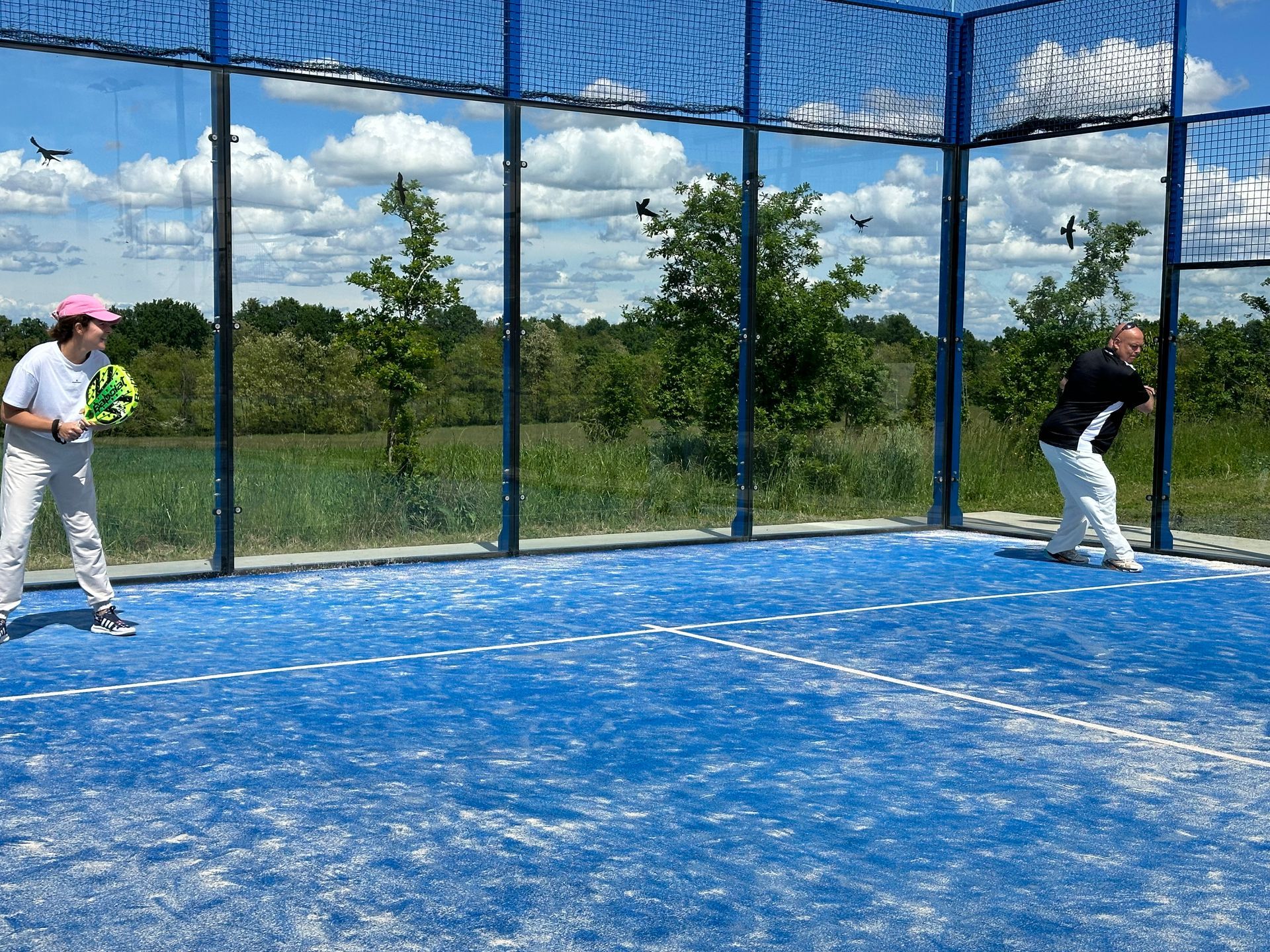 Due persone giocano a padel su un campo blu, recintato da una rete, sotto un cielo soleggiato.
