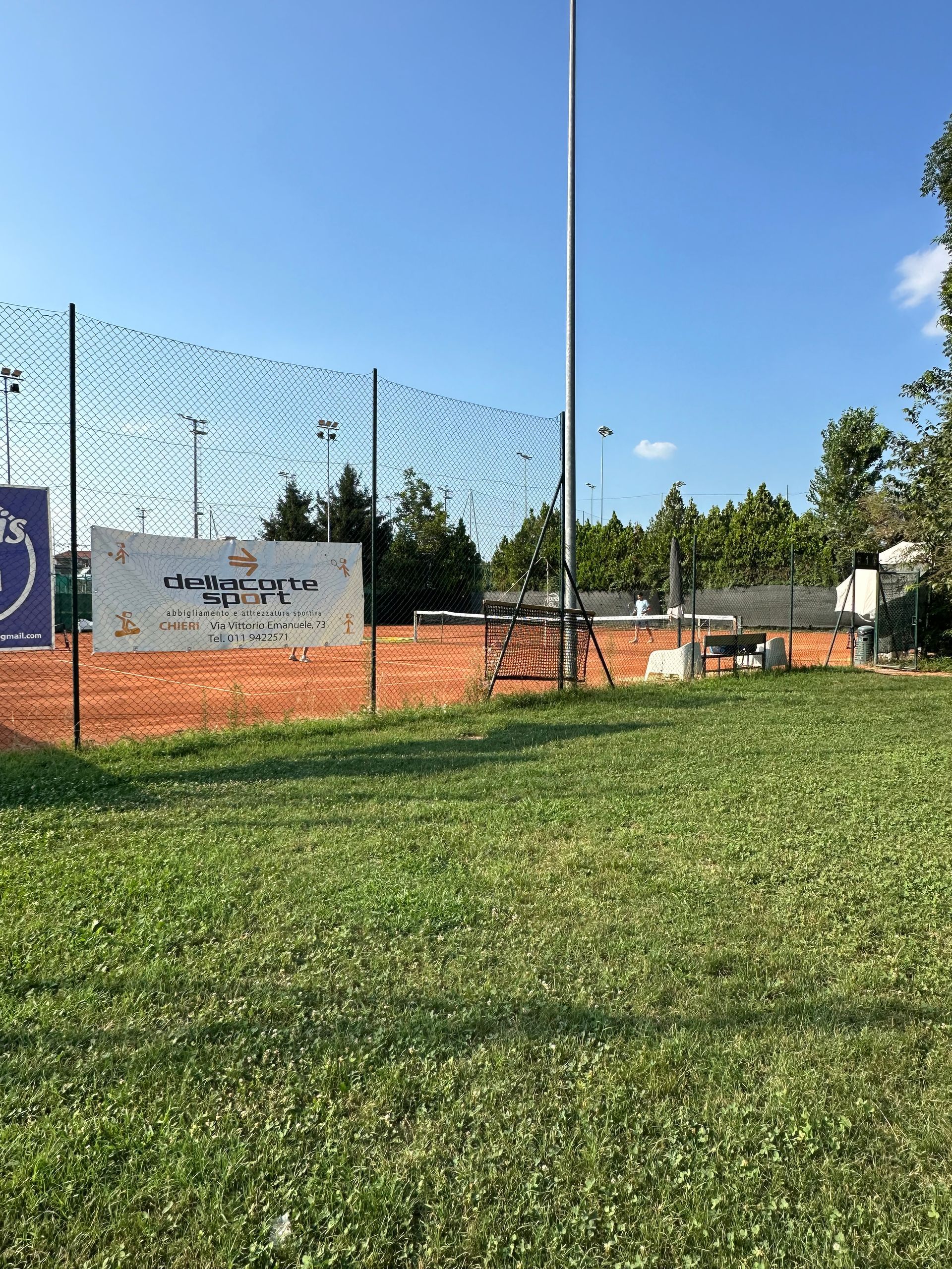 Campo verde con un campo da tennis dietro una recinzione. Un cartello è attaccato alla recinzione. Cielo azzurro.