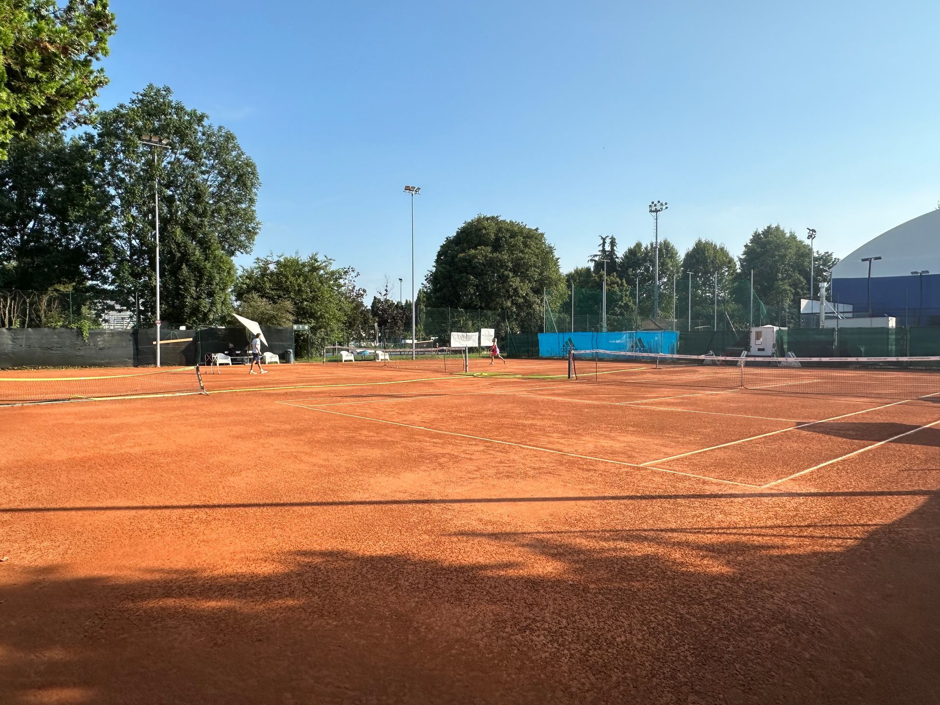 Campo da tennis in terra battuta sotto un cielo azzurro, circondato da alberi e luci alte.
