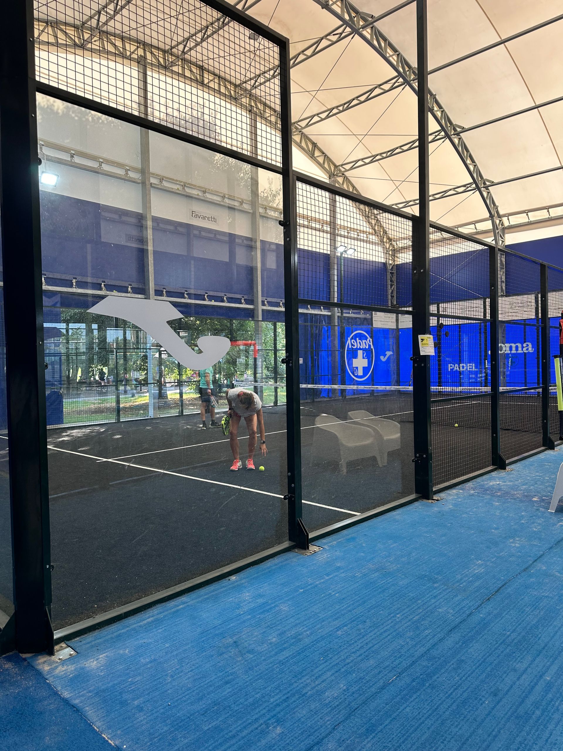 Campo da padel con superficie blu e pareti in rete nera. Una persona è in piedi al centro, gli altri giocatori sono visibili.