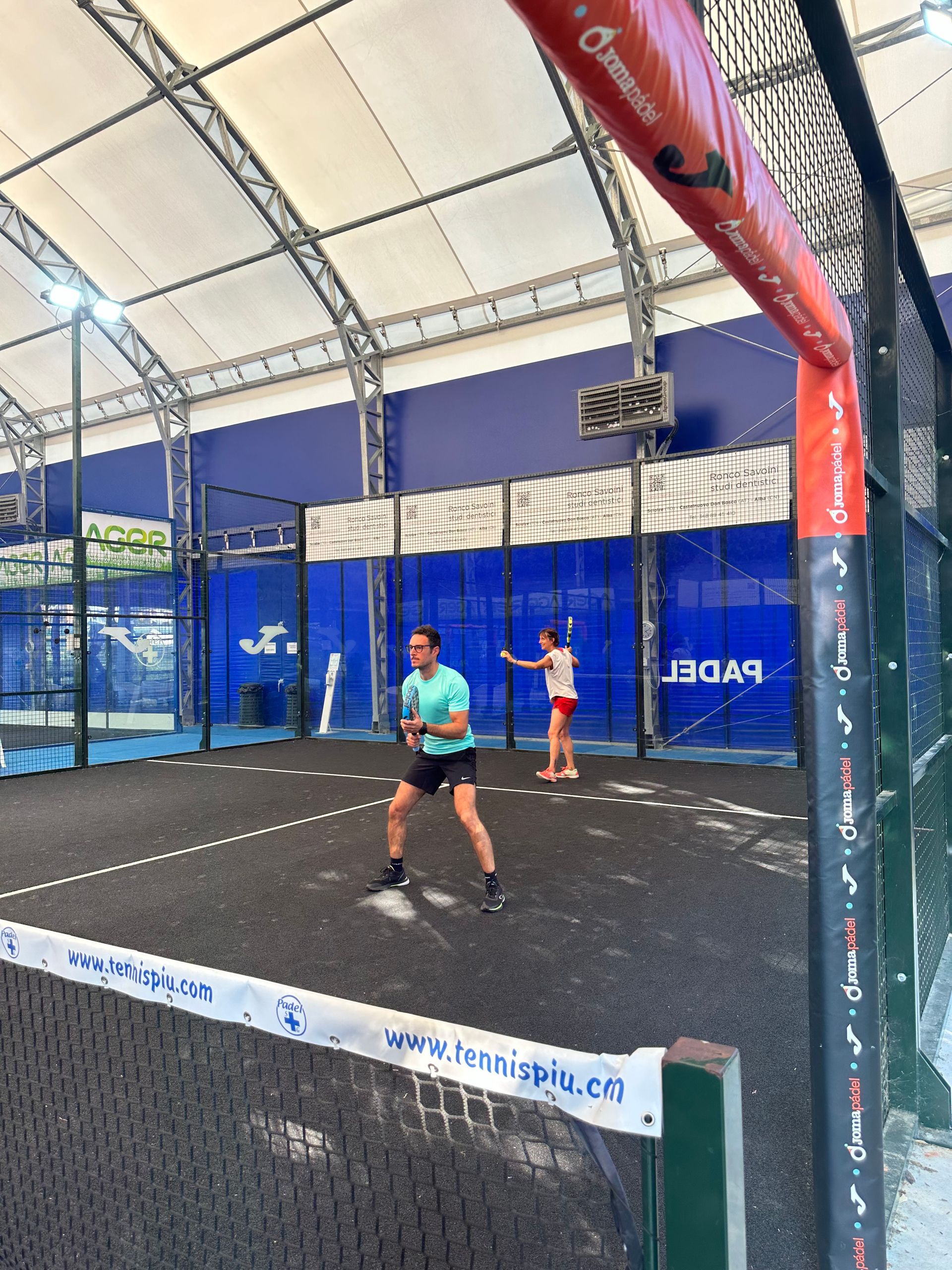Due persone giocano a padel su un campo con pareti blu e tetto bianco. Una persona sta per colpire la palla.