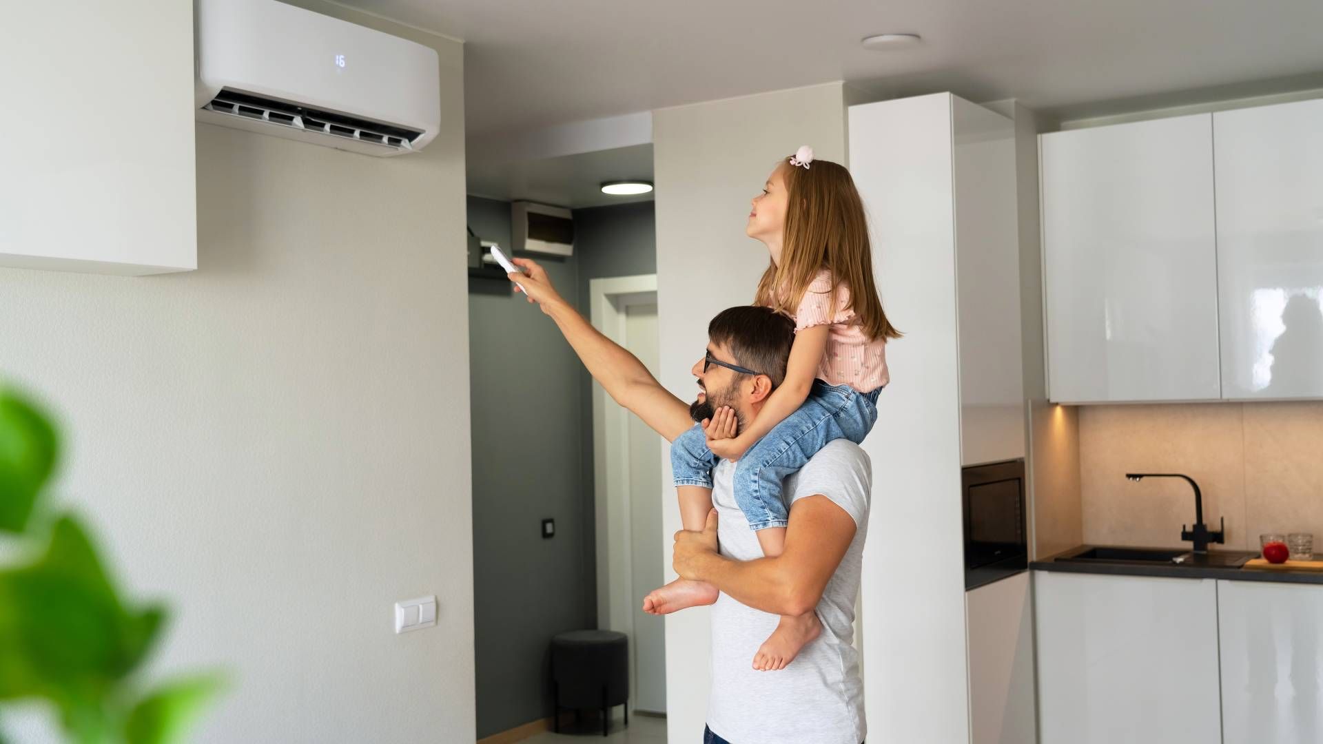 Man carrying a child on his shoulders while using a wall-mounted air conditioner remote in a modern apartment